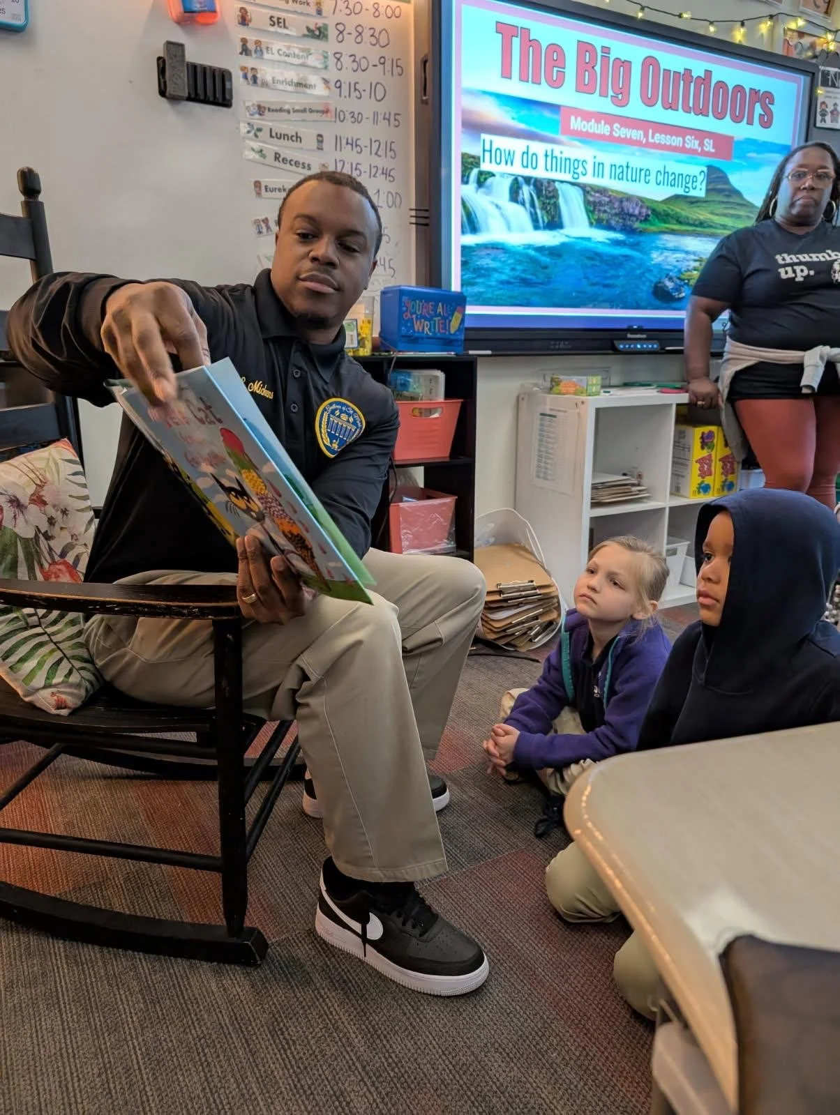 A man in a black shirt with a badge reading 'Marshall' reads a colorful children's book to two kids sitting on the floor in a classroom. A woman stands nearby, and a digital screen displays a lesson titled 'The Big Outdoors' with a question about how nature changes.