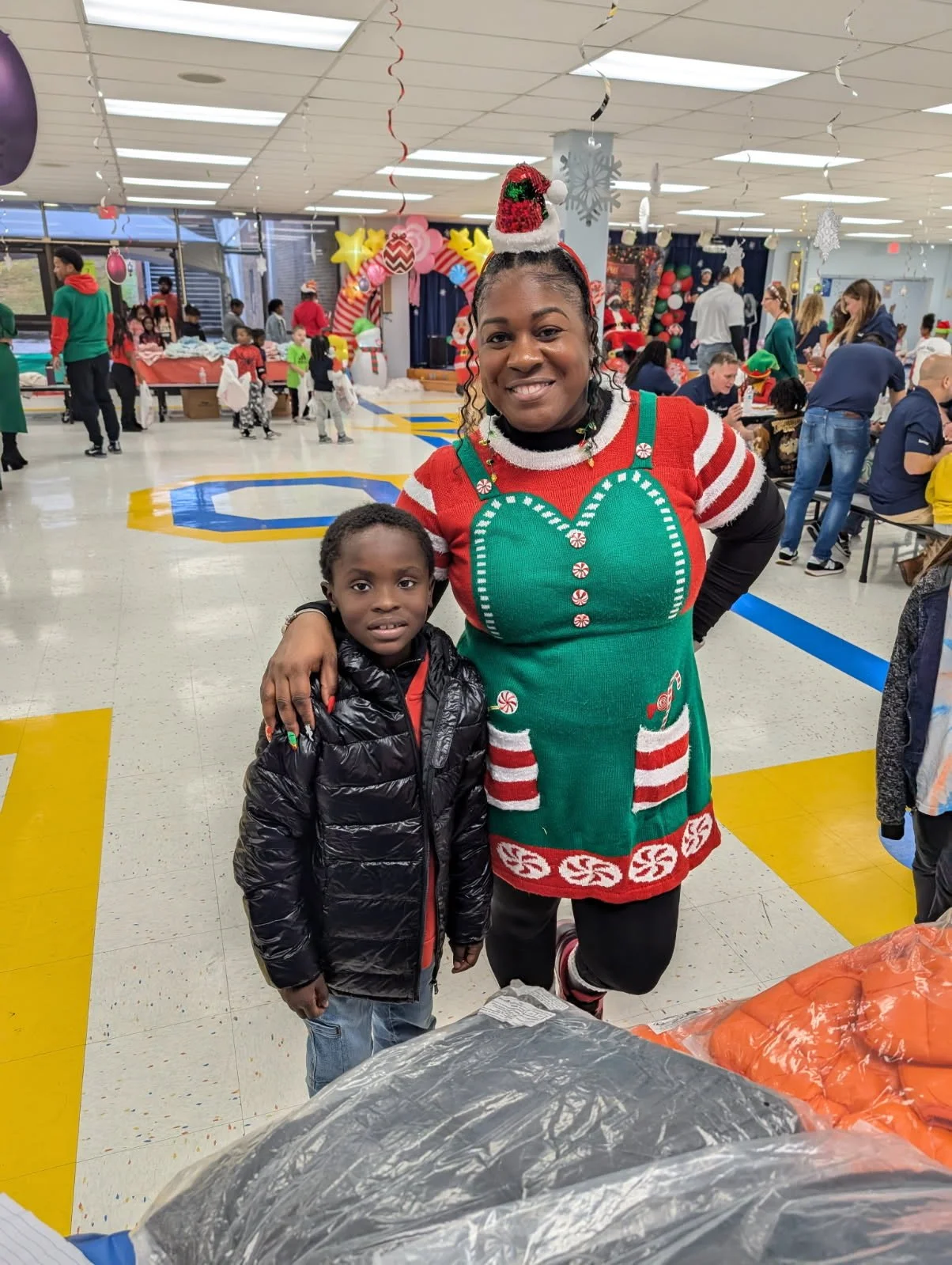 A woman dressed in a Christmas elf-themed sweater with holiday decorations, standing next to a young boy in a black puffy jacket at a festive indoor event with many people, Christmas decorations, and balloons in the background.