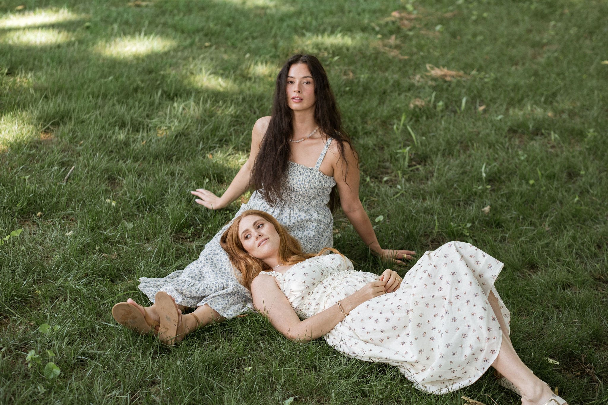 Two young women lying on the grass outdoors, one with long red hair resting her head on the other's lap and the other with long dark hair sitting nearby, both wearing summer dresses. Nashville Tennessee Women Female Model 