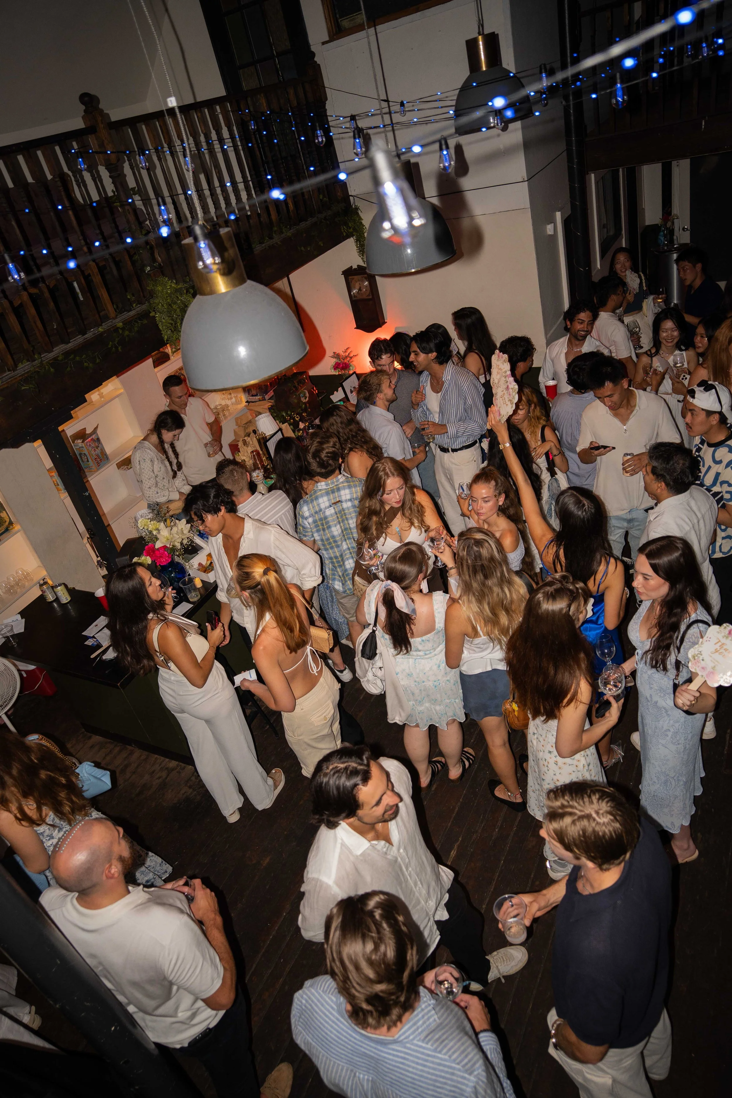 People gathered at a lively indoor party with string lights and large hanging lamps, socializing and holding drinks.