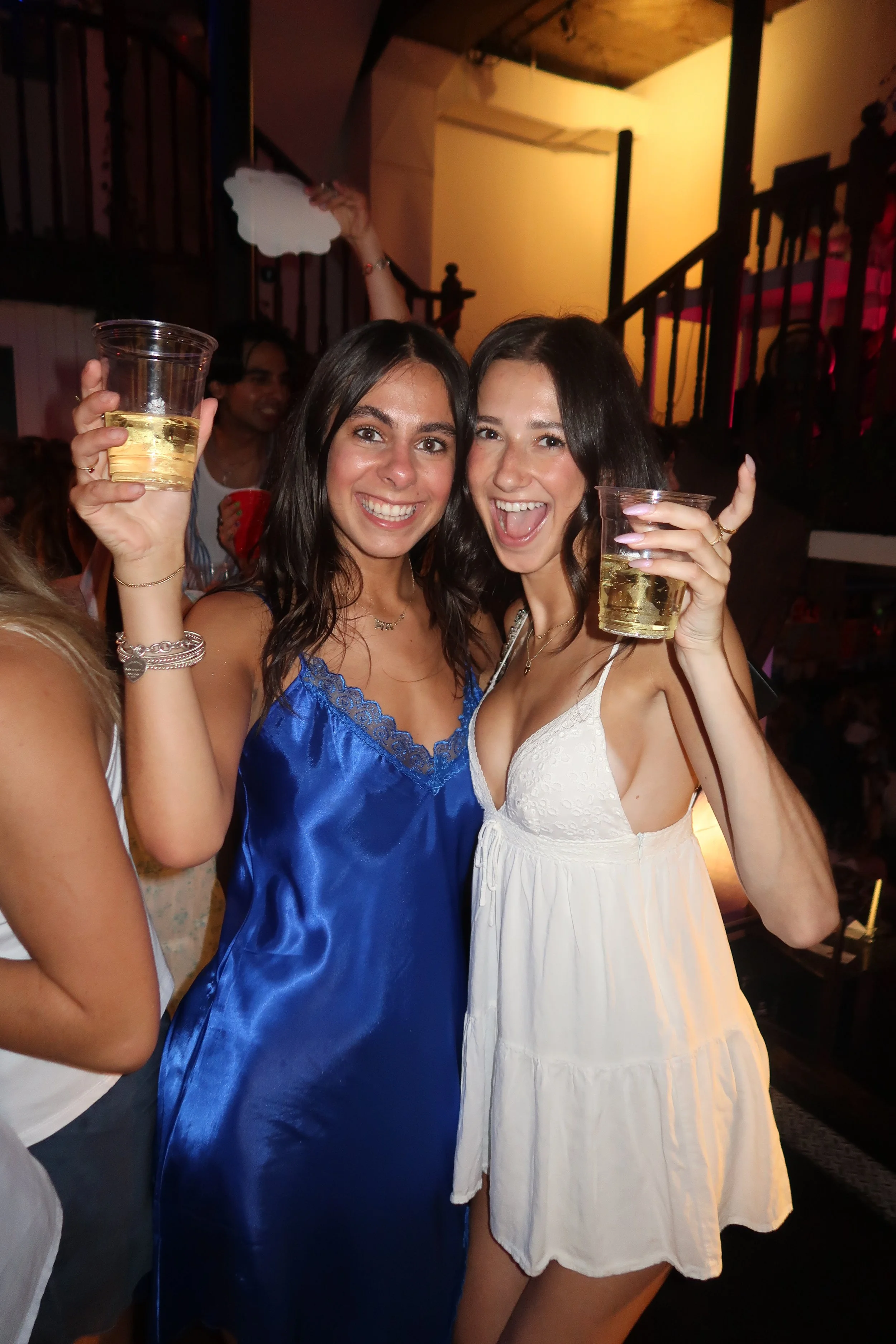 Two young women celebrating with drinks at a party, smiling and posing for the camera. One in a blue dress and the other in a white dress, holding plastic cups filled with a yellow beverage. Background features other people and party decorations.
