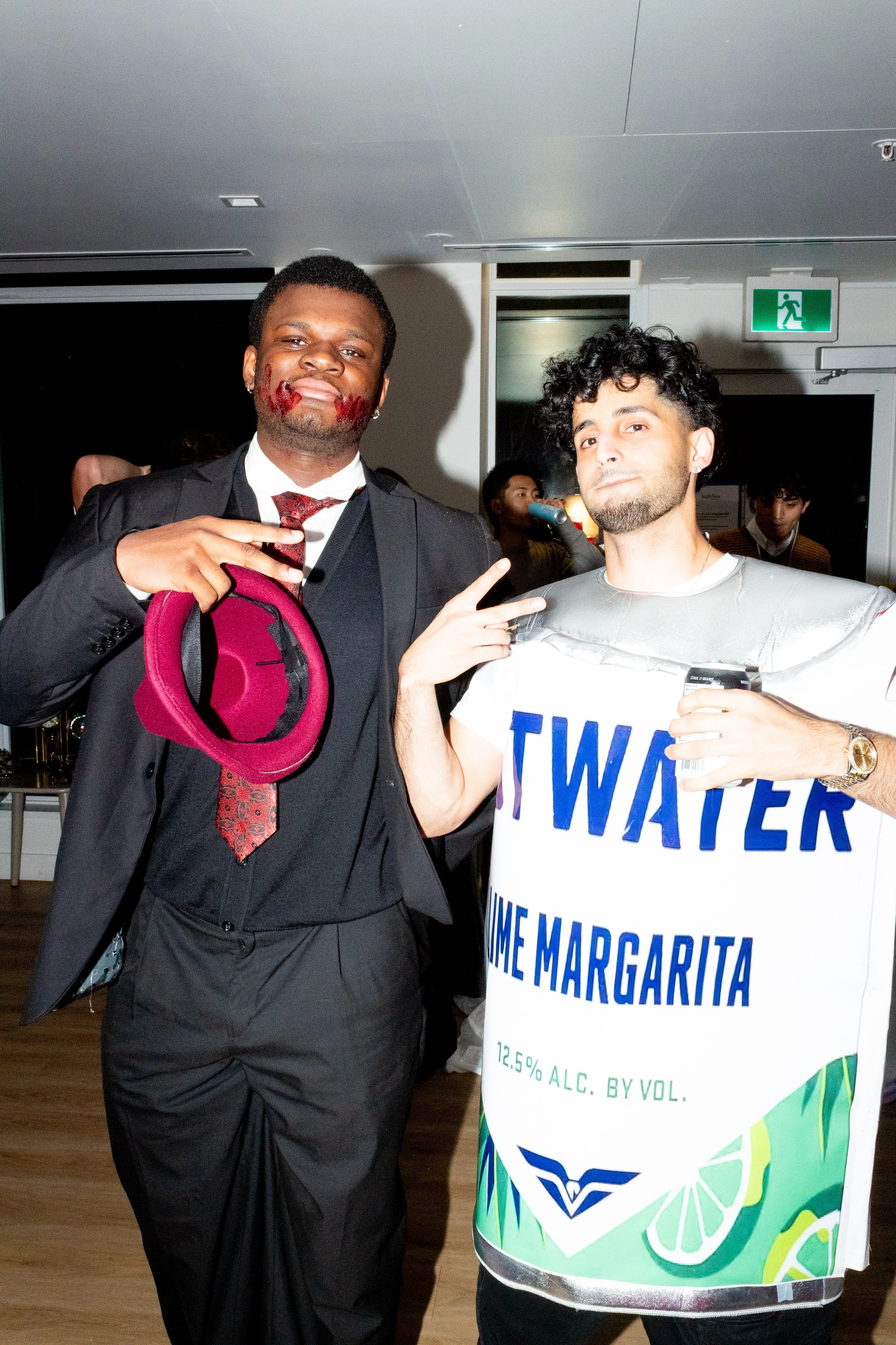 Two men dressed in costumes at a party. One man is wearing a tuxedo and holding a pink hat, with face paint that looks like blood on his lips. The other man is dressed as a juice carton labeled "Twat" with a Margarita flavor, holding a drink and maki
