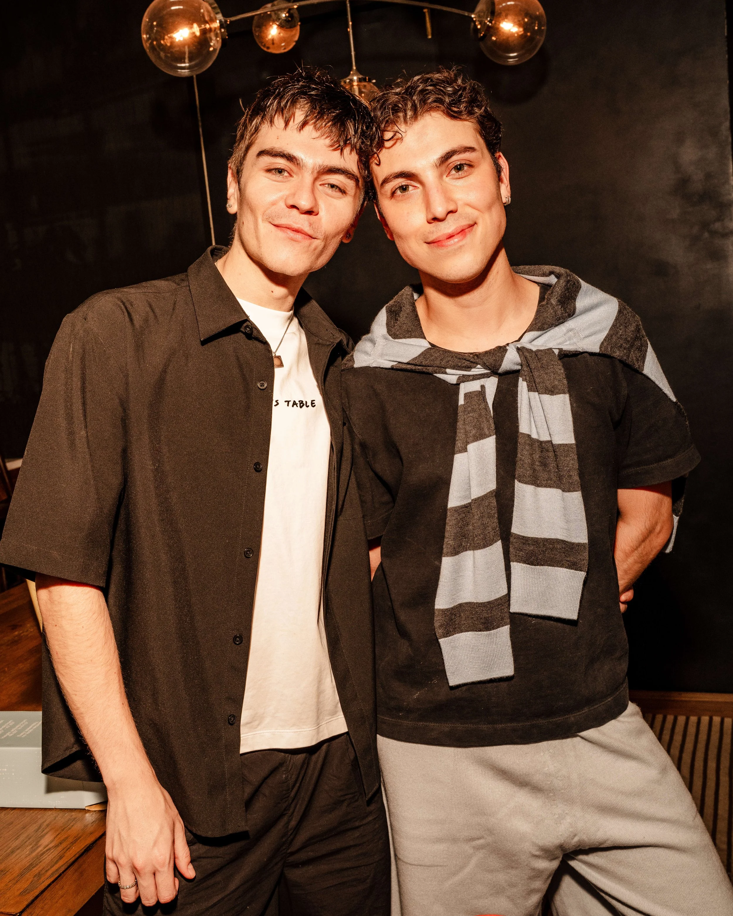 Two young men standing close together, smiling, in a dimly lit room with black walls and hanging decorative globe lights.