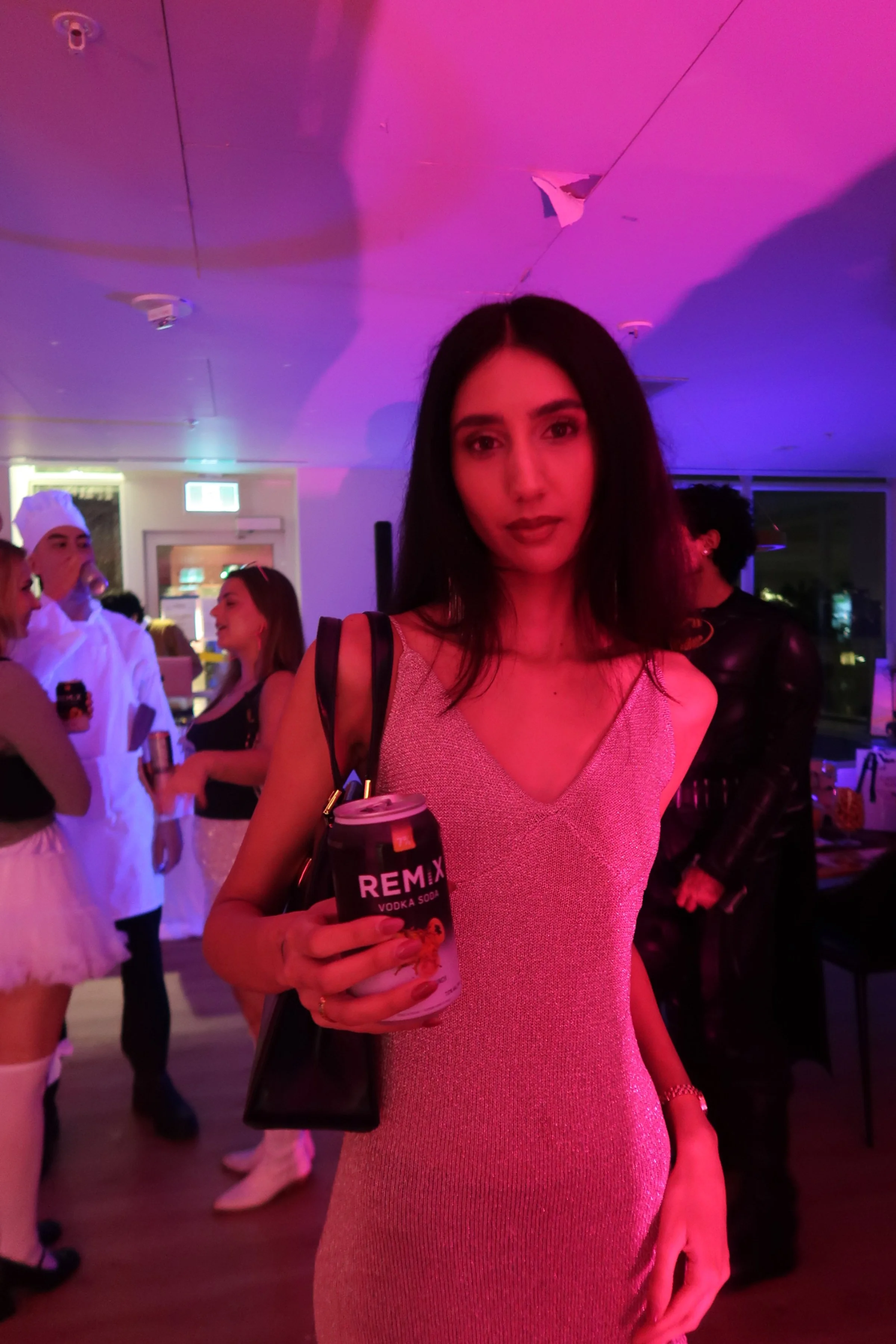 A woman in a sparkly pink sleeveless dress holding a can of REMIX vodka soda at a party or social gathering, with other people in costumes and festive attire in the background under purple and pink lighting.