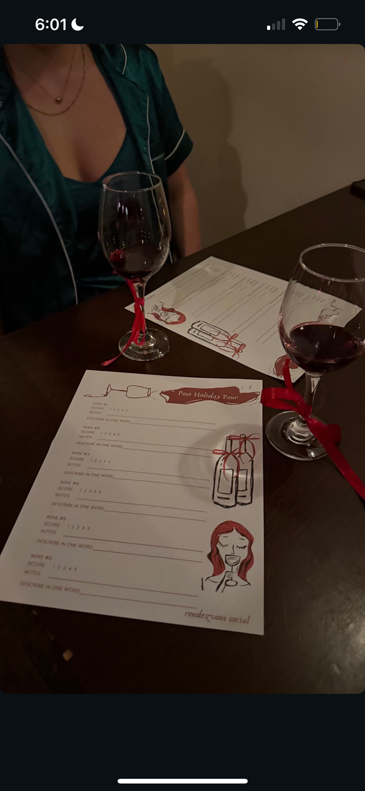 Two glasses of red wine with red ribbons tied around the stems, on a table with wine tasting score sheets, one with a cartoon woman and wine bottles, and a woman wearing a green satin shirt in the background.