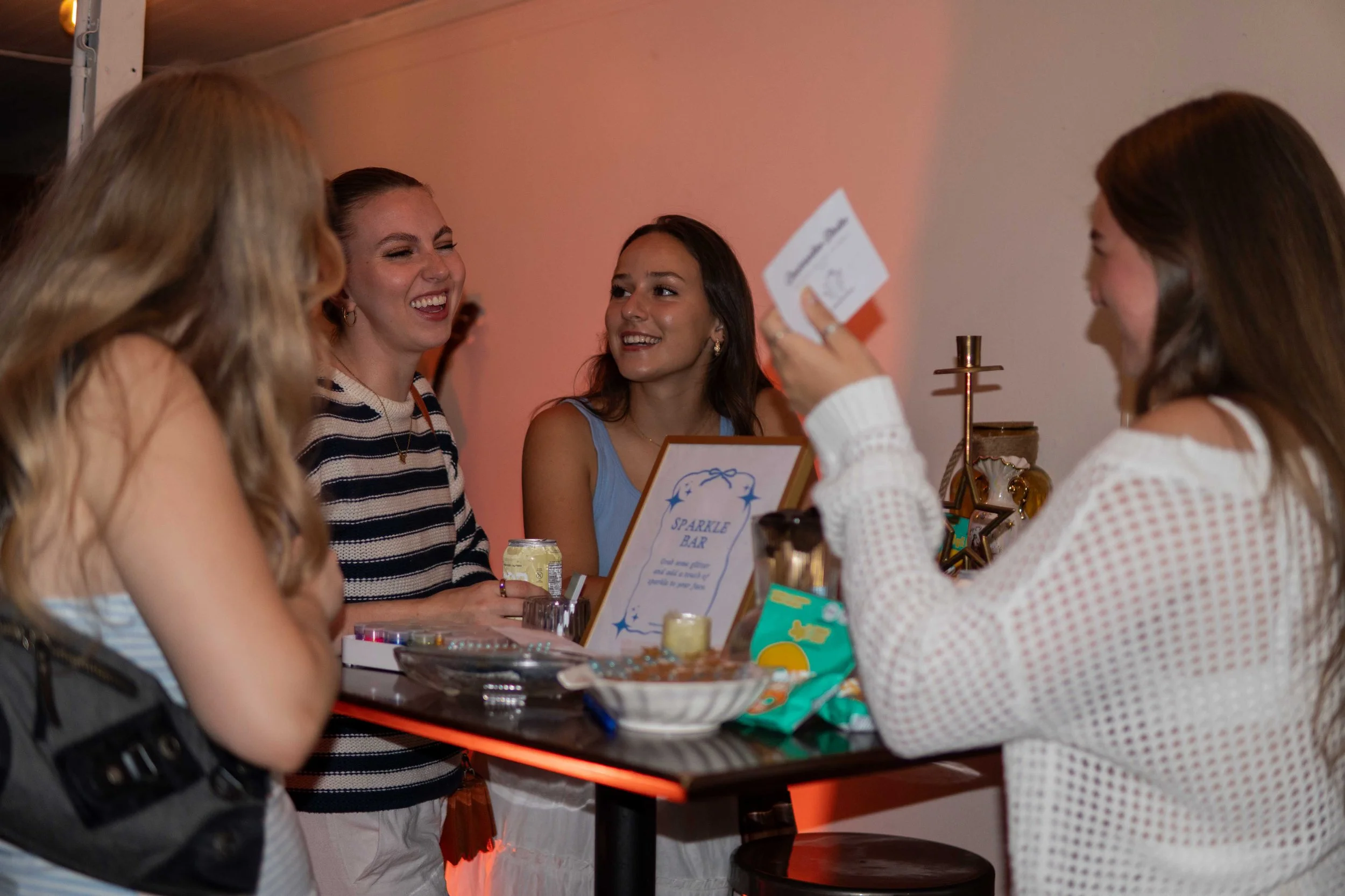 Four women are gathered around a bar counter, smiling and talking at a party or event.
