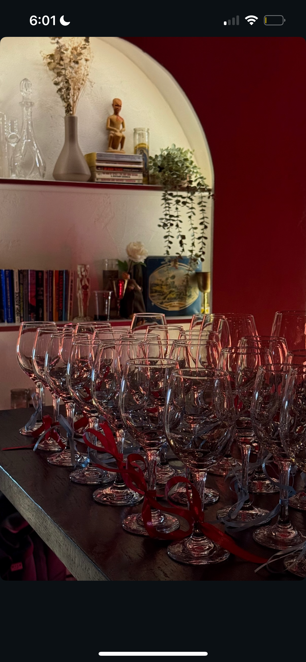 Multiple empty wine glasses with red and silver ribbons on a wooden table, with shelves holding books, plants, and decorative items in the background.