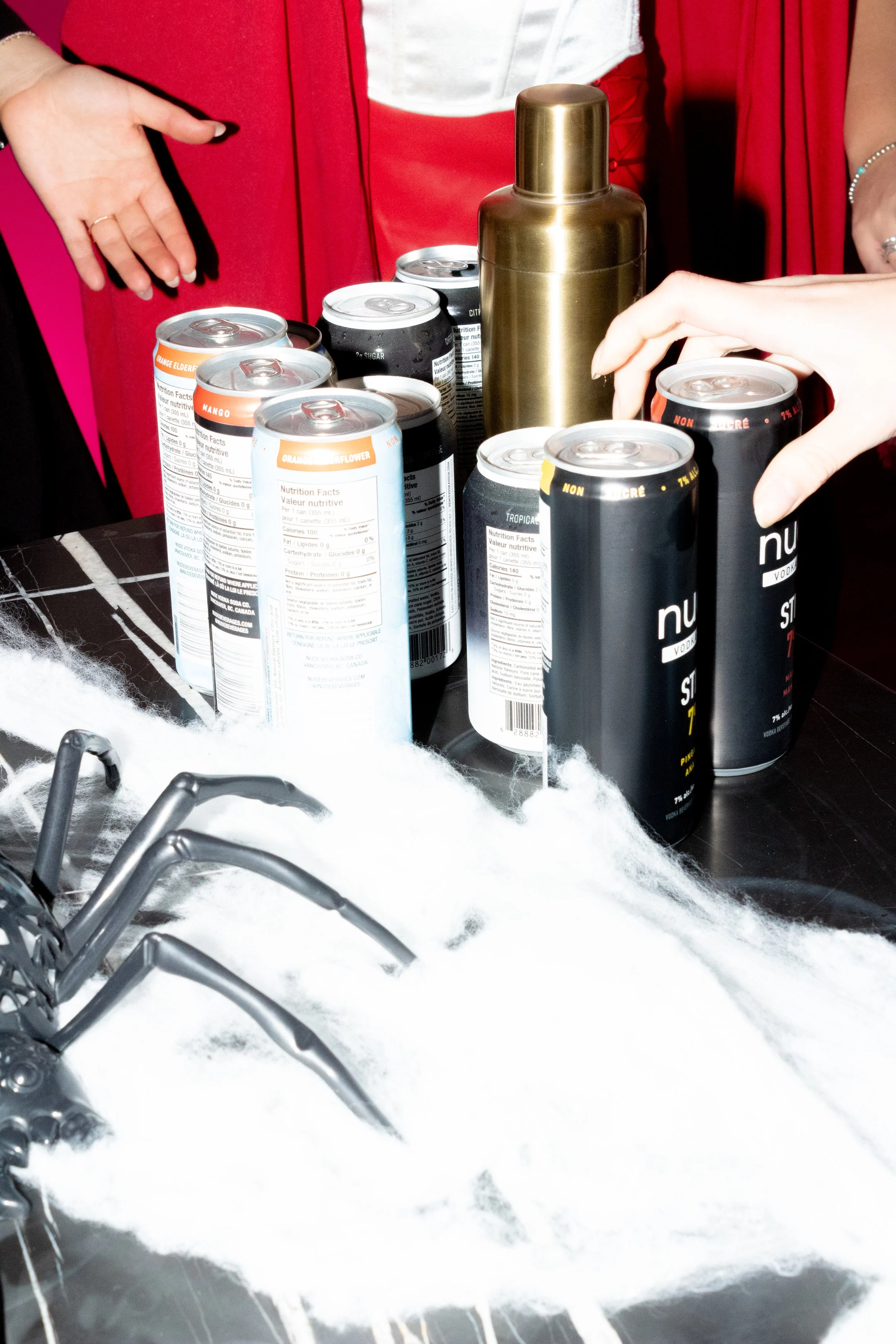 A Halloween-themed table with cans of soda, a gold thermos, and synthetic spider web on a black surface.
