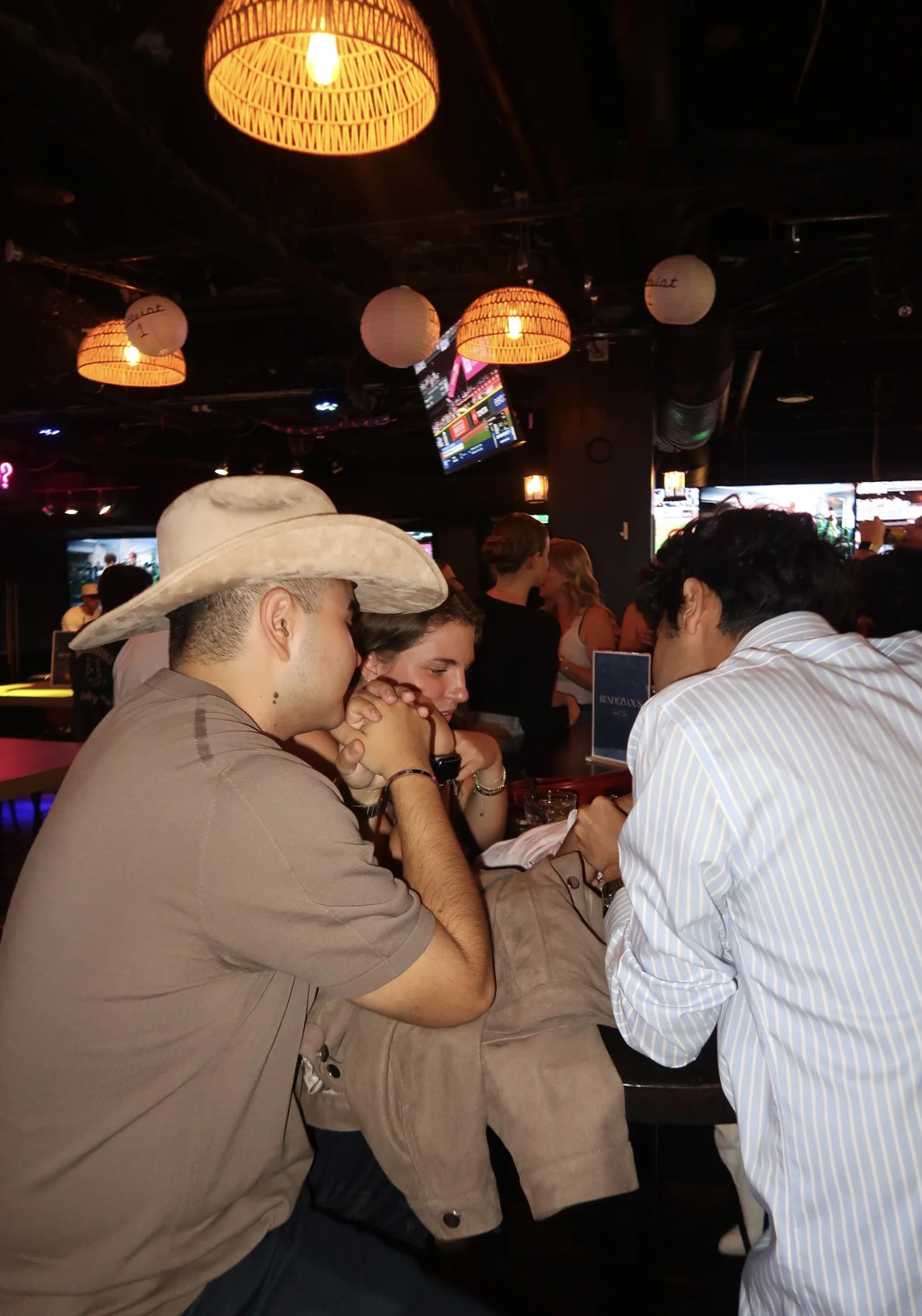 Three young adults sitting at a bar in a dimly lit room, engaged in conversation, with hanging lamps overhead and multiple screens displaying sports or news in the background.