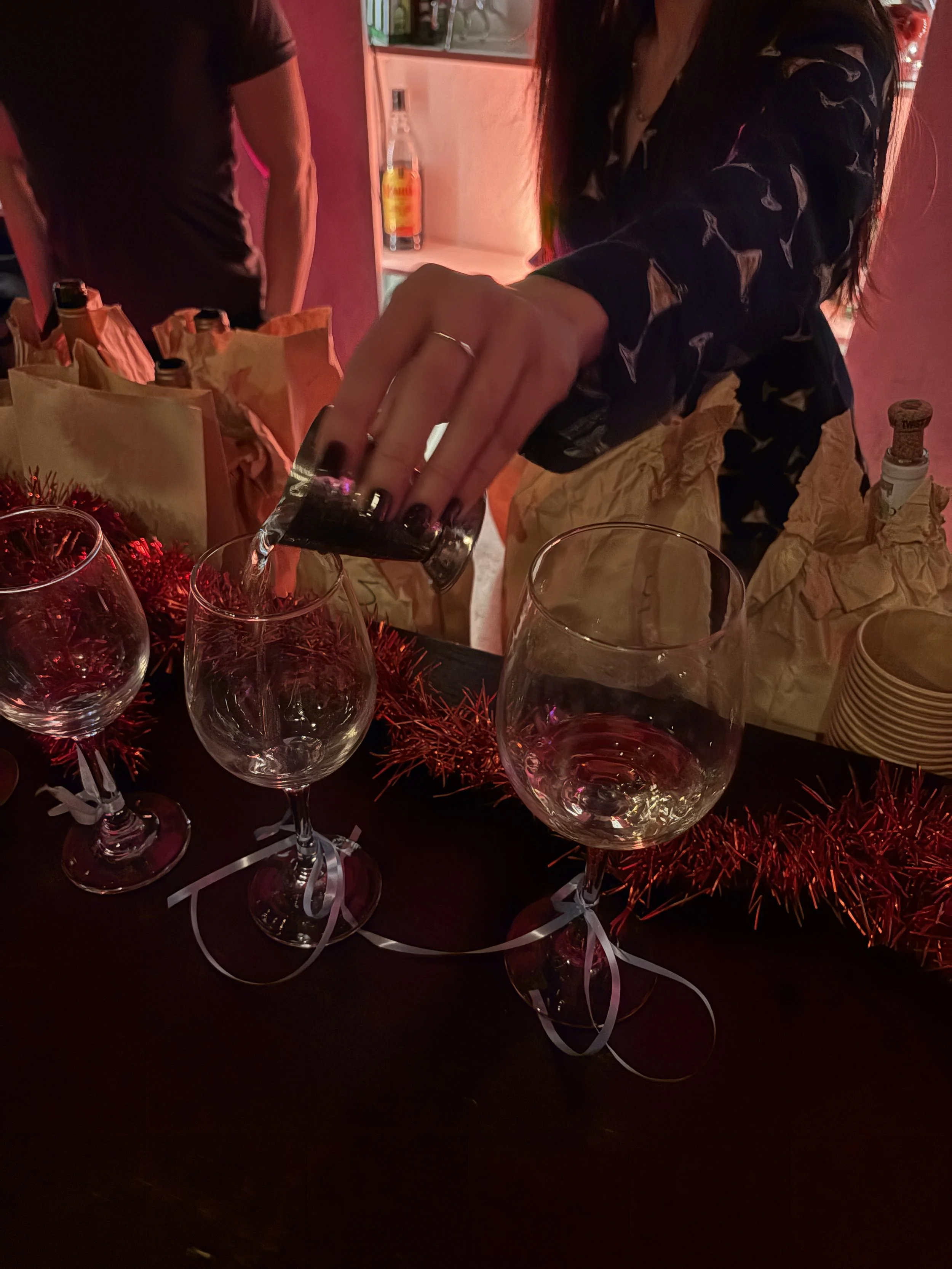 Person pouring clear liquid into a wine glass at a celebration or party, with other wine glasses and party decorations on the table.