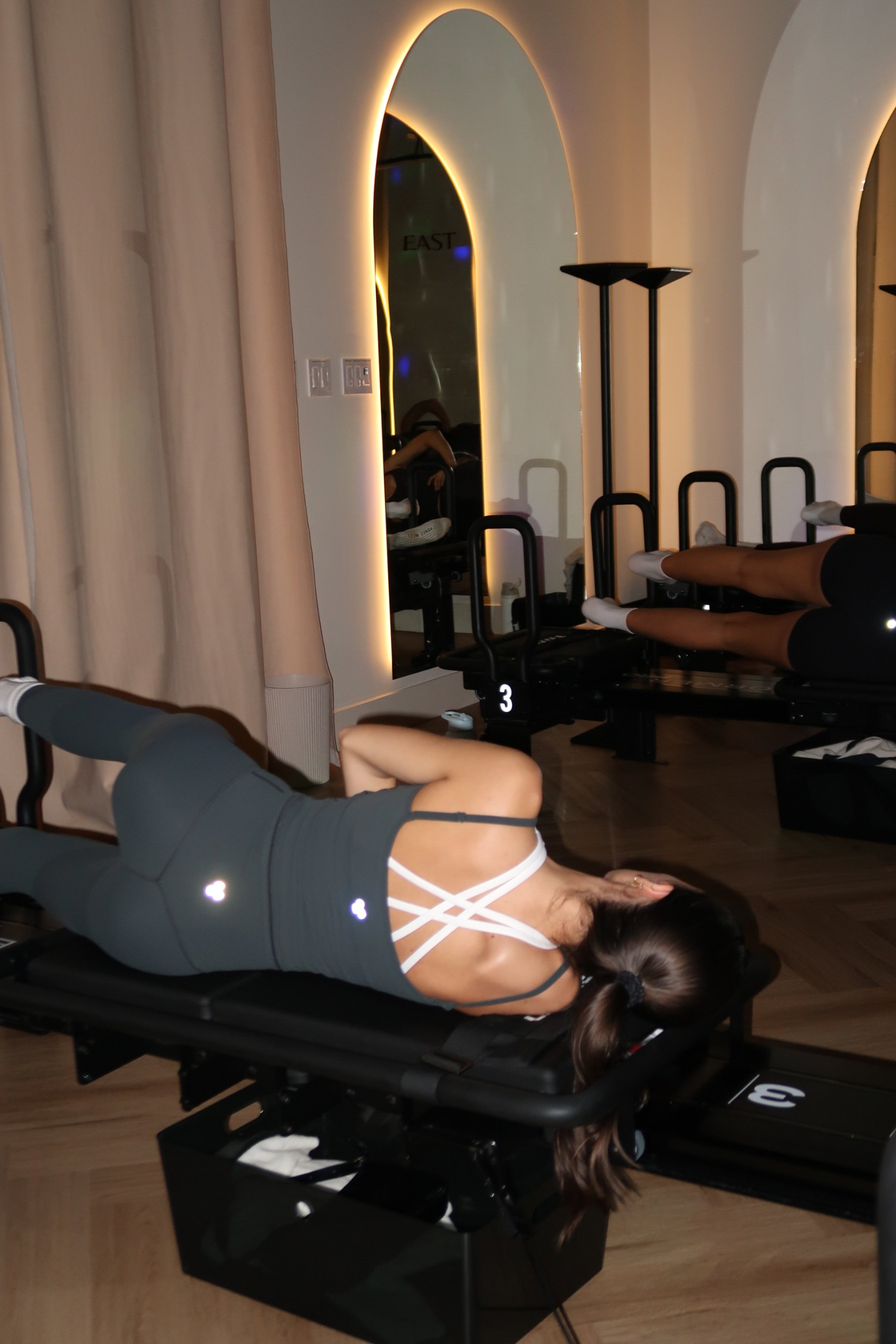 Woman lying on a Pilates reformer machine in a fitness studio, with mirrors and other exercise equipment in the background.
