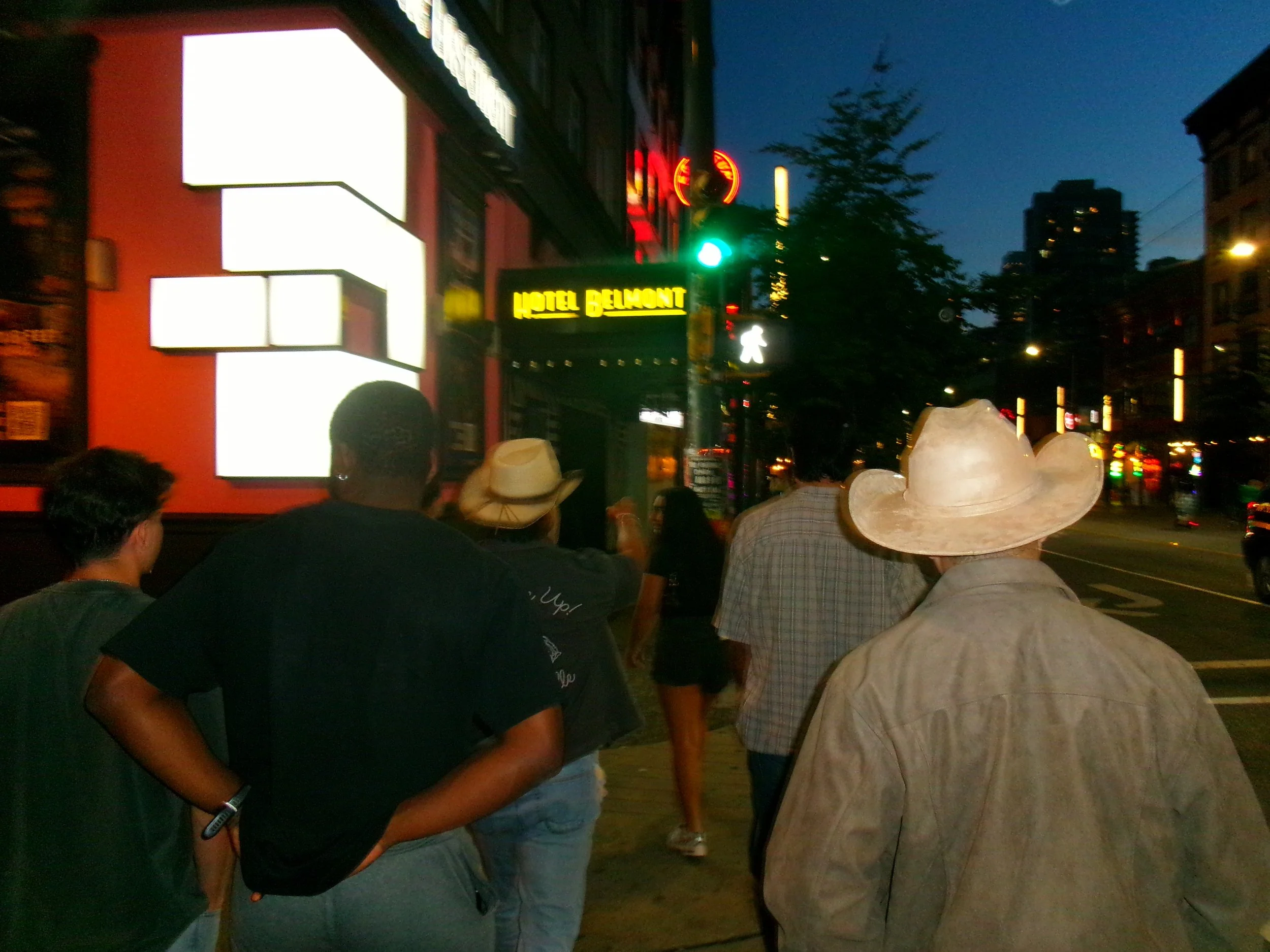 People walking on a city sidewalk at night with a neon sign reading 'HOTEL BELMONT' visible in the background.