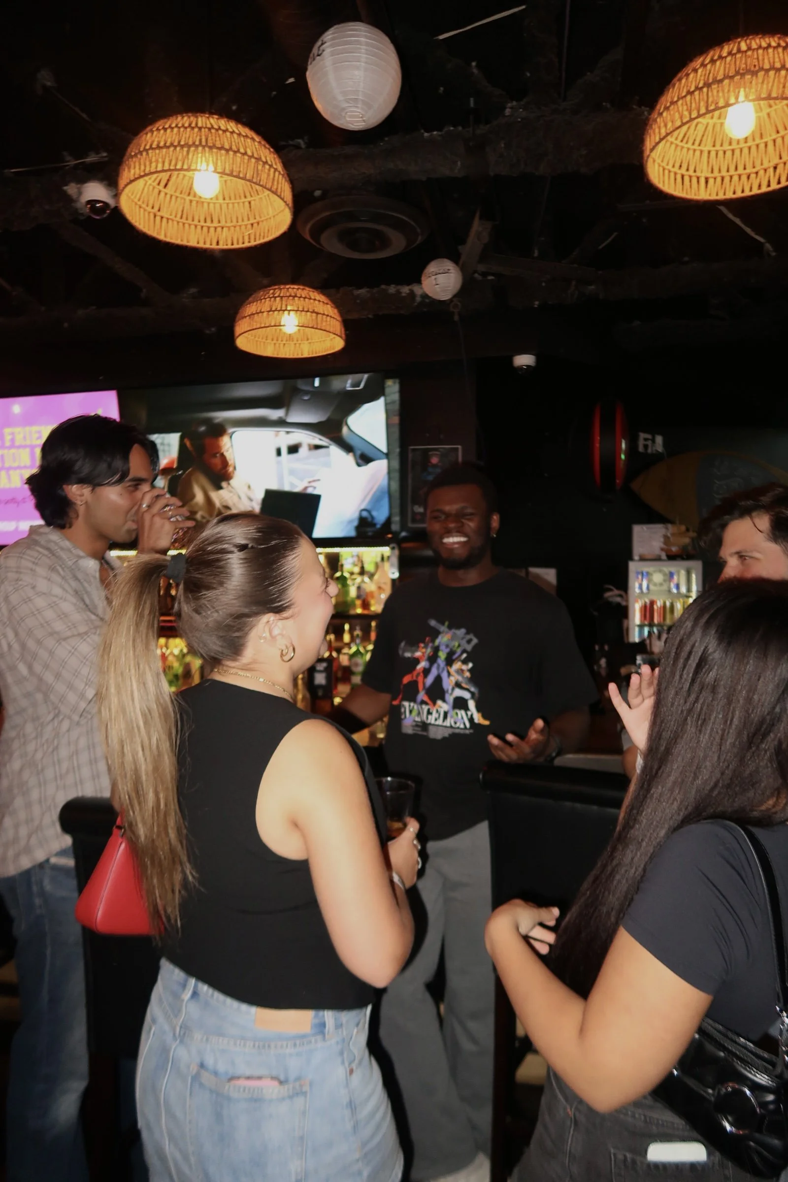 Group of friends socializing at a bar with warm lighting and a television screen in the background.