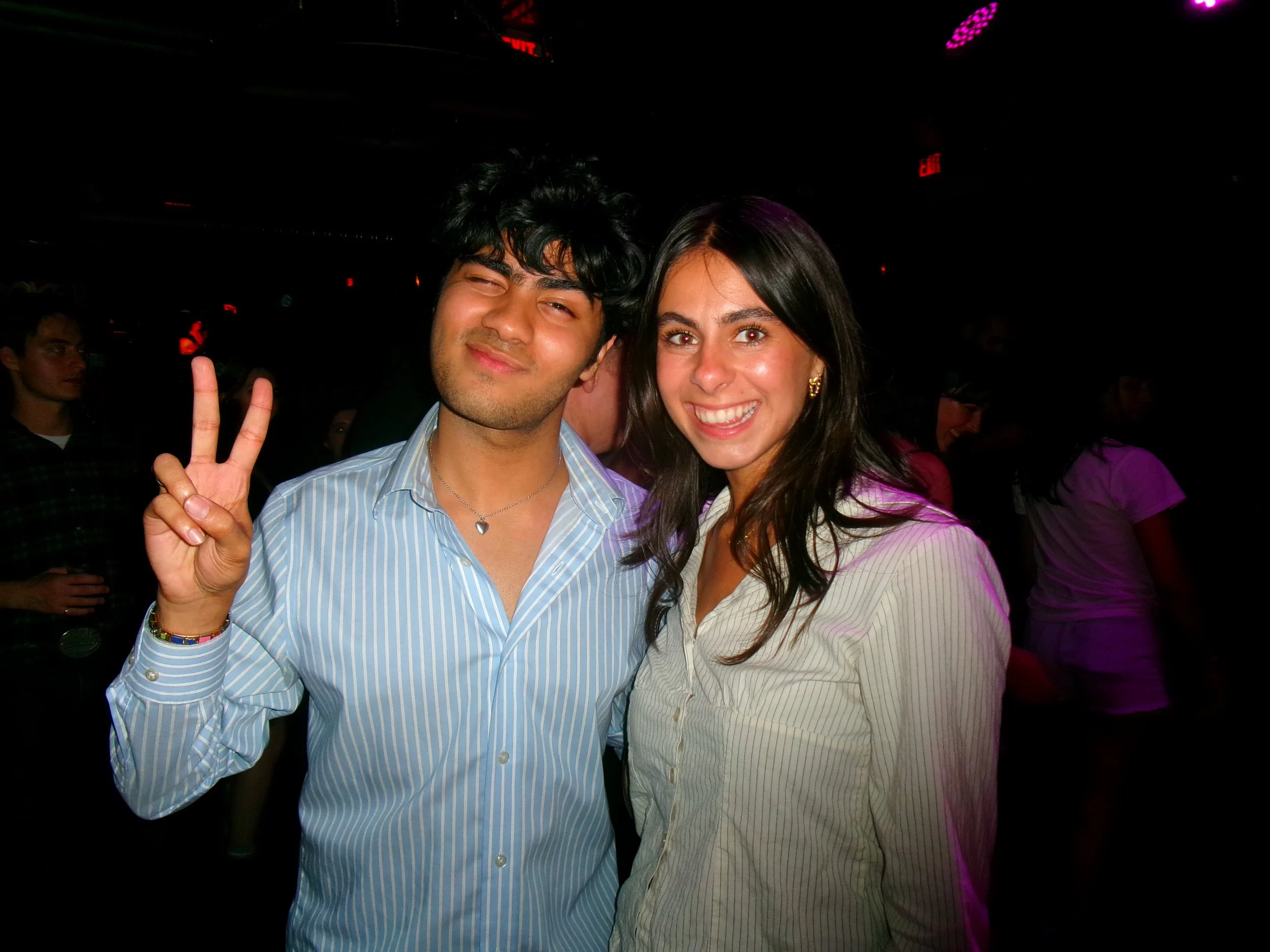 Smiling young man making a peace sign with her fingers, standing next to a smiling young woman, both in casual shirts, at a lively indoor event or party.