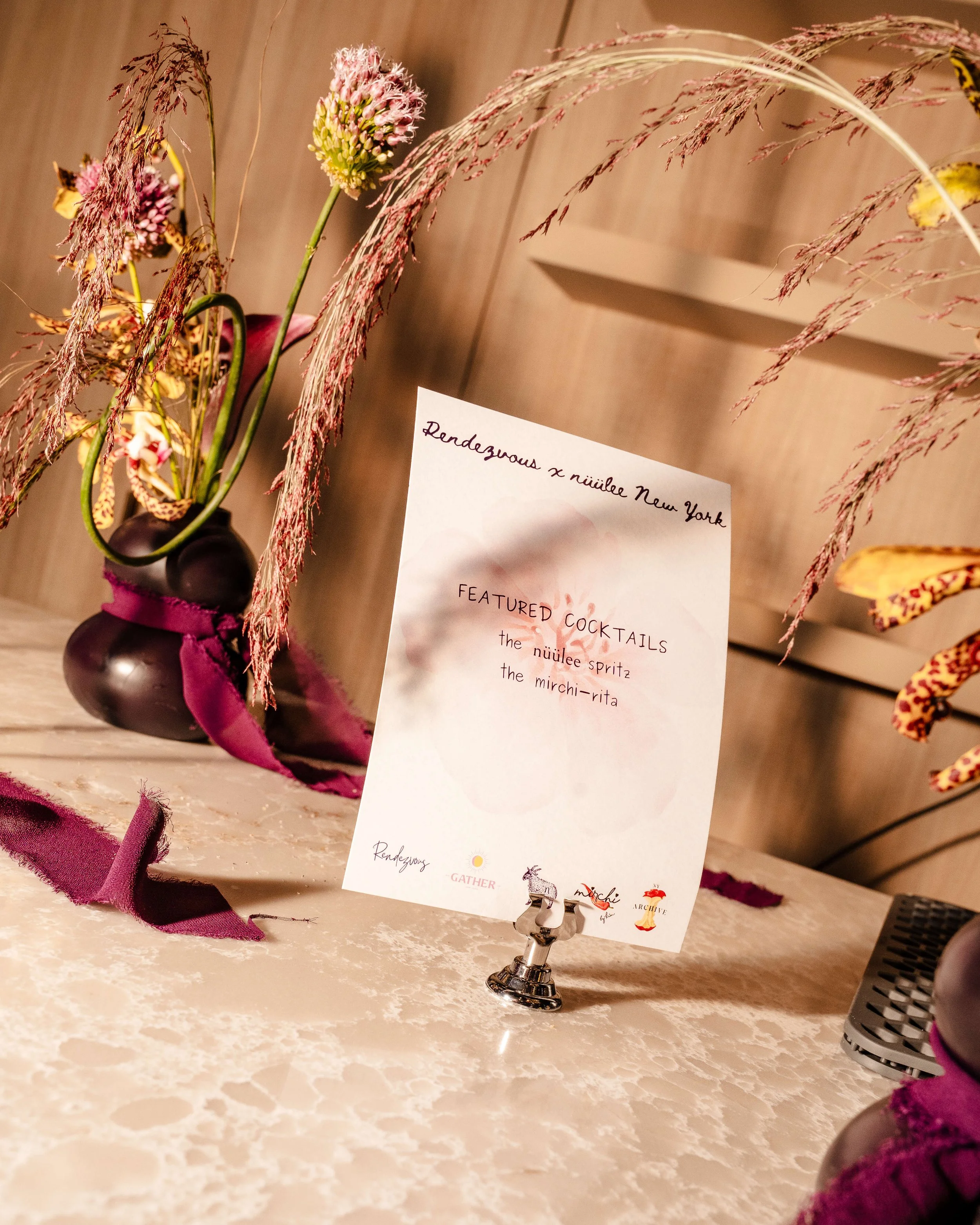 A cocktail menu on a table at a fancy event, listing featured cocktails: the nuuile spritz and the mirchi-rita, with floral decorations and a dark glass vase with flowers.