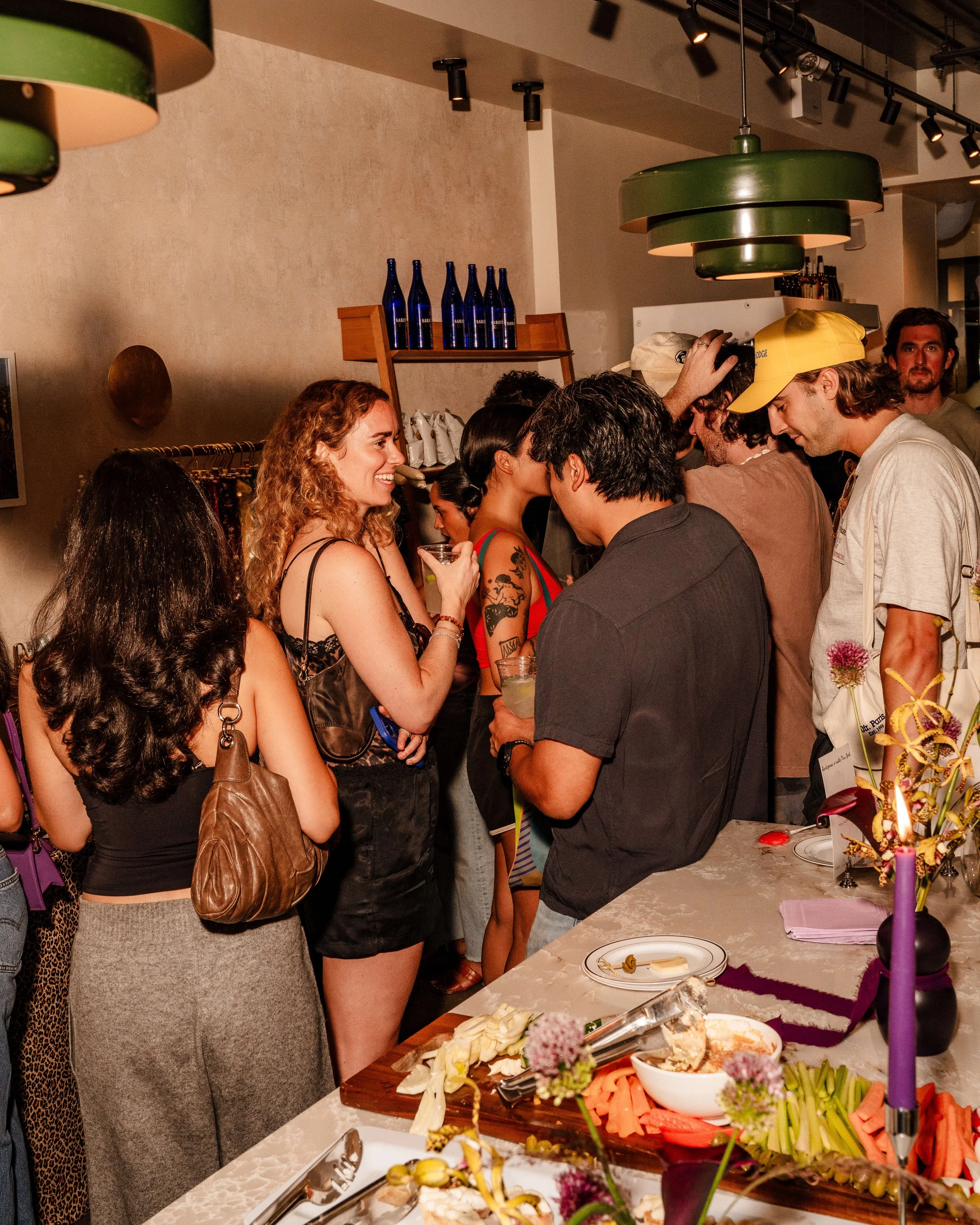 People mingling at a social gathering, standing near a buffet table with food and drinks in a lively indoor setting.