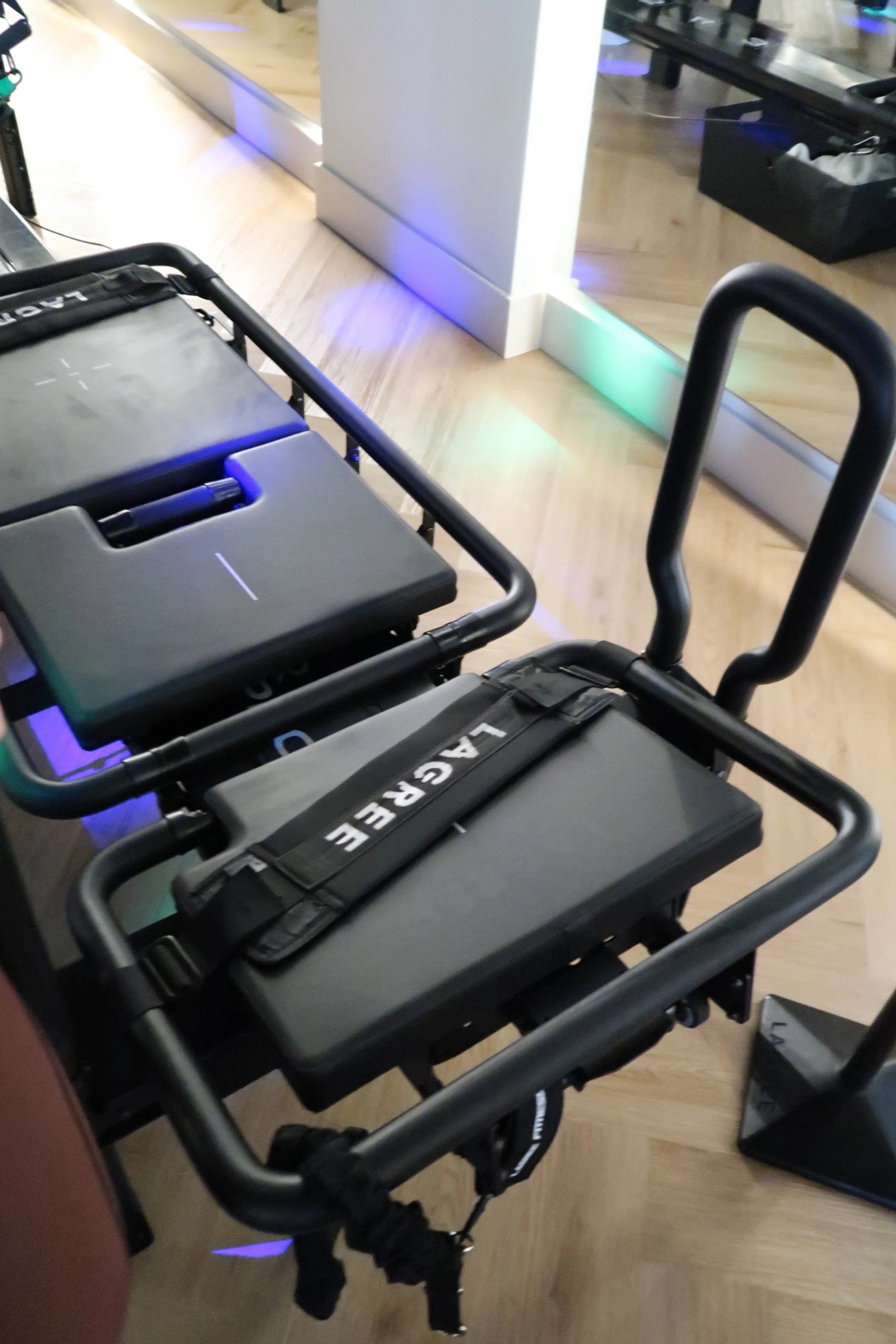 Two airport luggage carts with black straps labeled 'LAGRE' in an indoor setting, near a glass barrier with colored lights reflected on the floor.