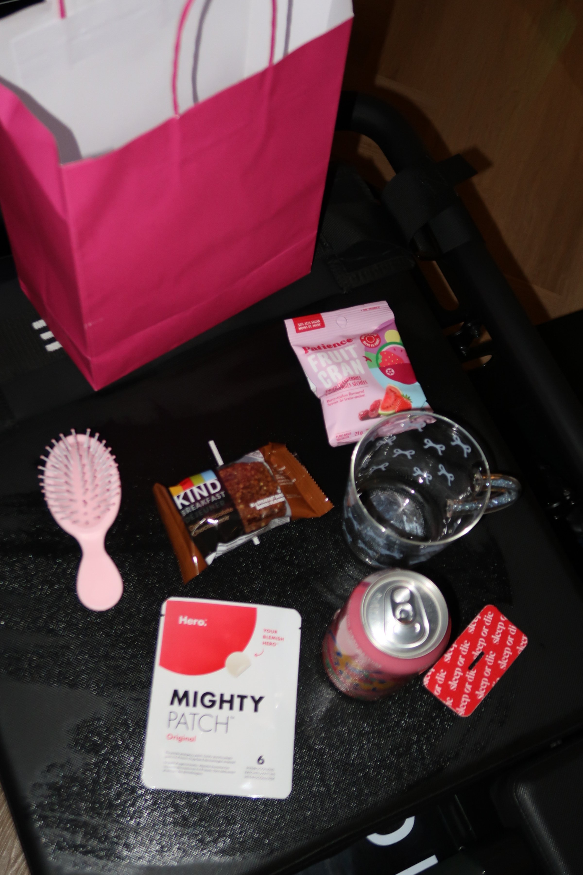 A pink gift bag, a pink hairbrush, a pack of Kind breakfast bars, a packet of fruit cranberry snacks, a glass of water, a soda can, and a pack of Mighty Patch acne patches on a black surface.