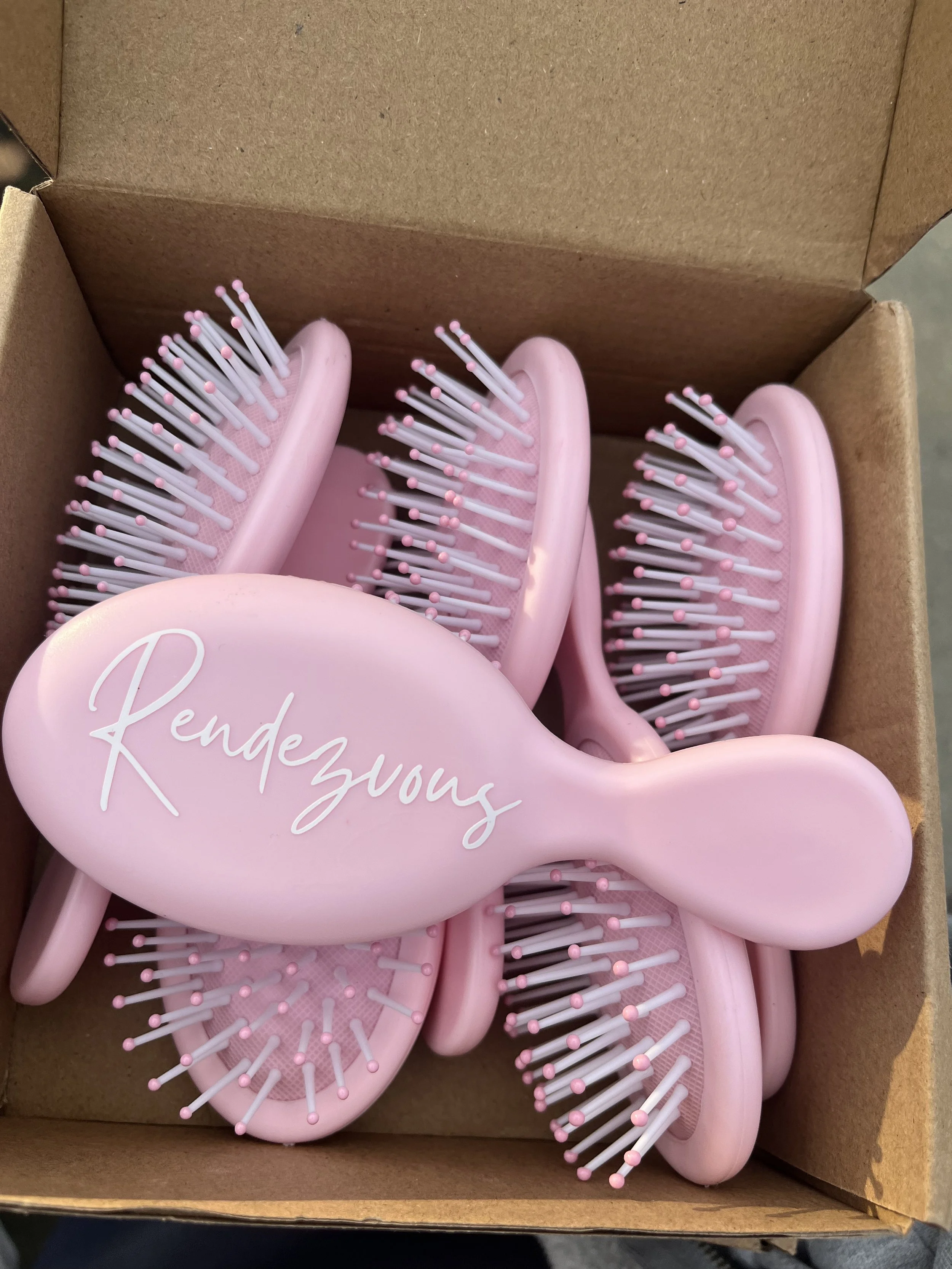 Close-up of a pink hairbrush with the word 'Rendezvous' written on it, inside with several similar pink hairbrushes in a cardboard box.