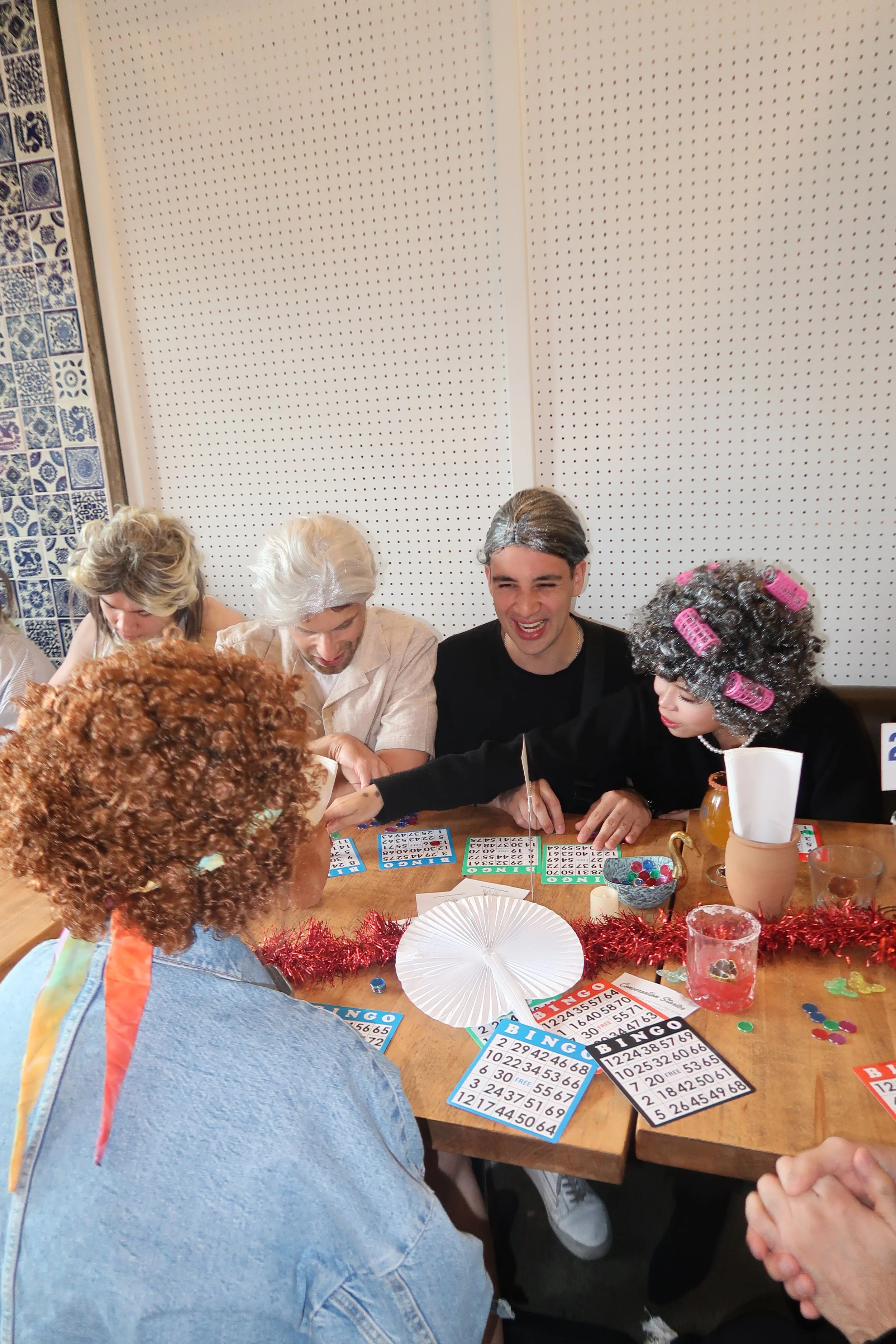 People dressed as old-fashioned seniors and a person in a wig with curlers play bingo at a decorated table.