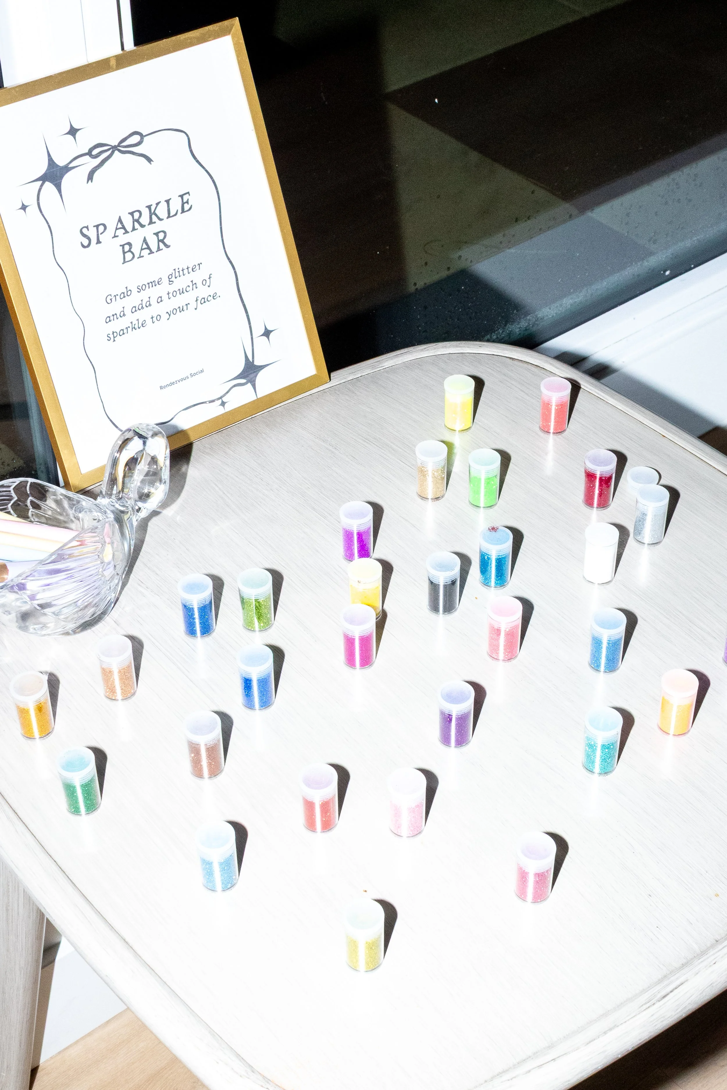 Table with small containers of multicolored glitter, a sign that reads 'Sparkle Bar,' and a glass swan figurine.
