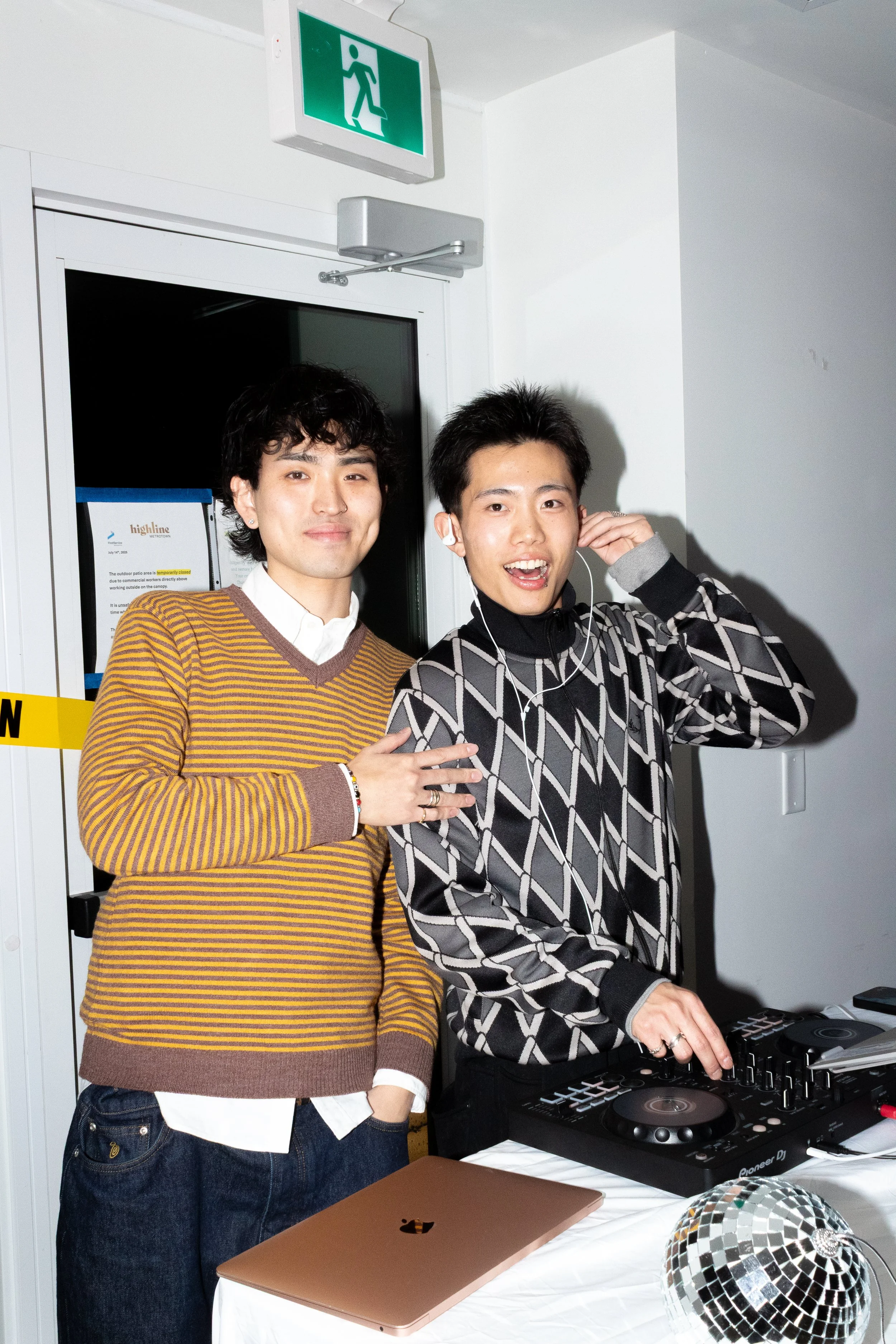 Two young men standing indoors; one is wearing a mustard yellow and brown striped sweater, the other is wearing a black and gray geometric patterned hoodie. The man in the hoodie is DJing with a Pioneer DJ controller, while the other has his hand on 