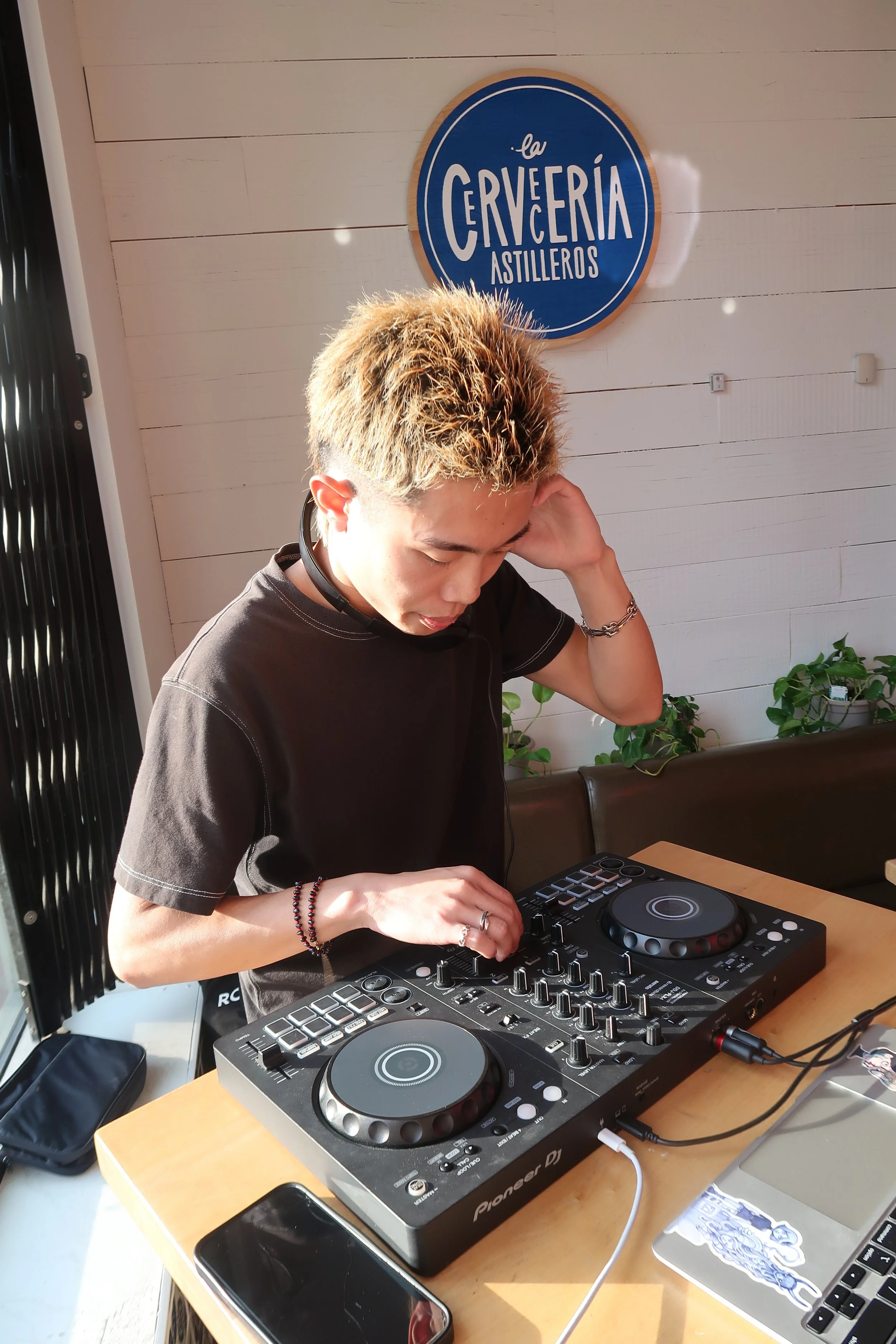 A young person with blonde hair uses DJ equipment at a table, with a sign reading 'La Cerveza Astilleros' on the wall behind them.