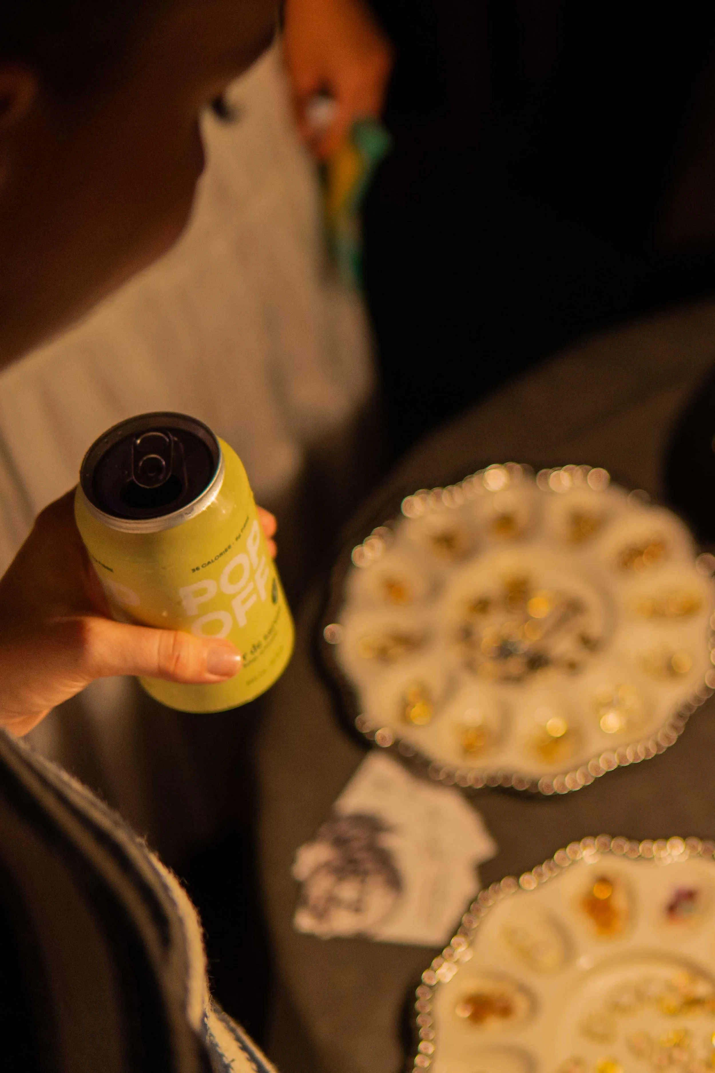 Person holding a yellow can of Pop Off with a tab opening, on a table with plates of snack foods and a napkin with a black and white design, in a dimly lit setting.