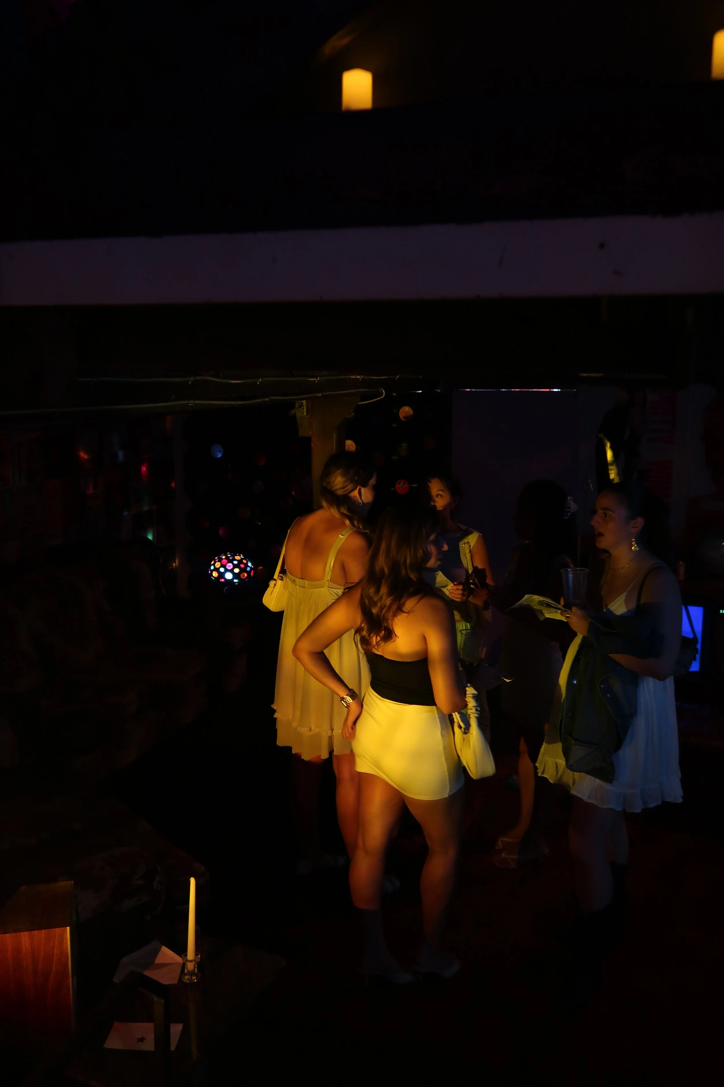 A group of women in yellow and black dresses are in a dimly lit bar or club, engaged in conversation. They are surrounded by faint decorative lights and a dark background.