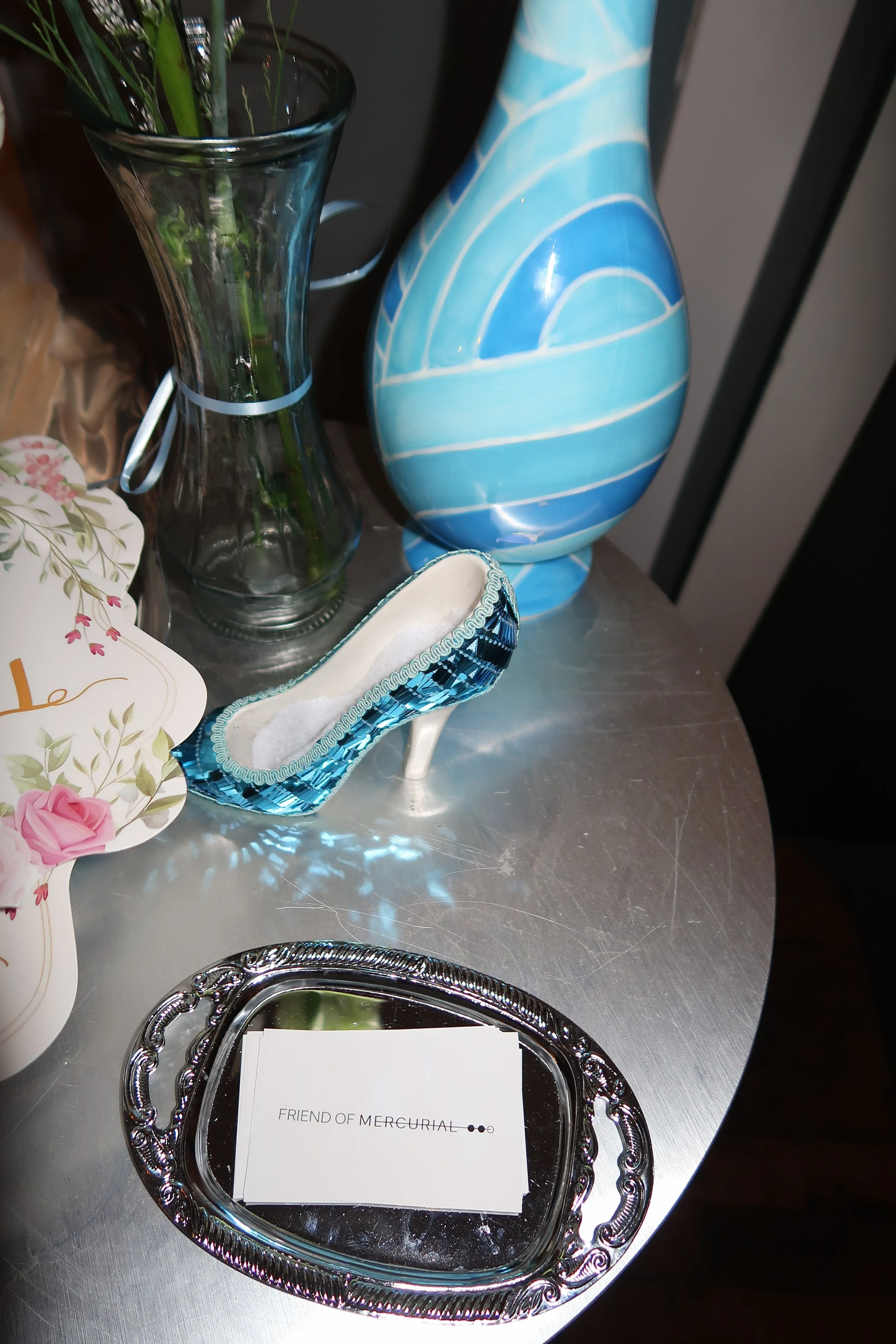 A silver tray with the text 'Friend of Mercurial' placed on a round metallic table. On the table, there is a porcelain shoe-shaped object, a glass vase with green plant stems, and a blue and white decorative vase. Part of a pink floral card and a whi