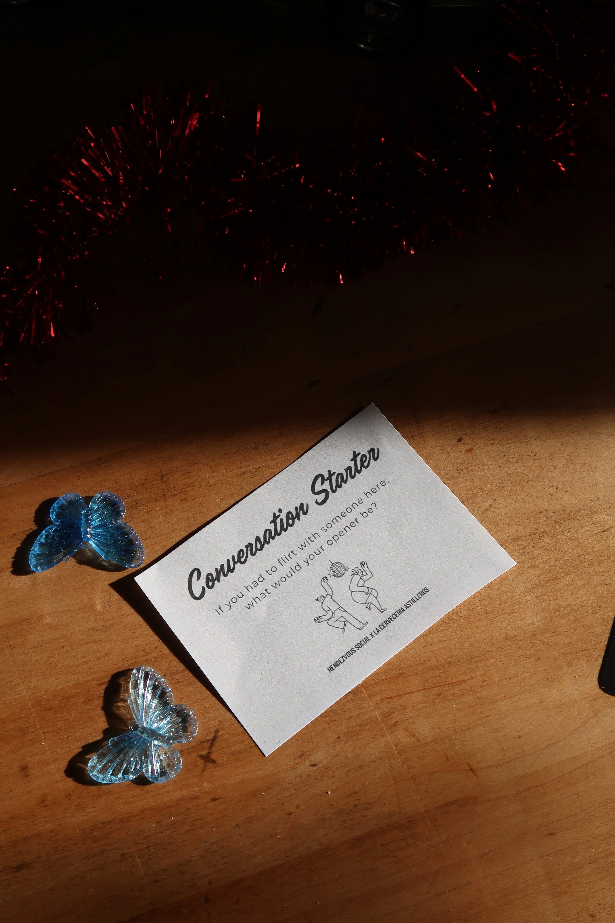 A conversation starter card on a wooden table with decorative blue butterflies and red tinsel in the background. The card asks, 'If you had to flirt with someone here, what would you open be?'.