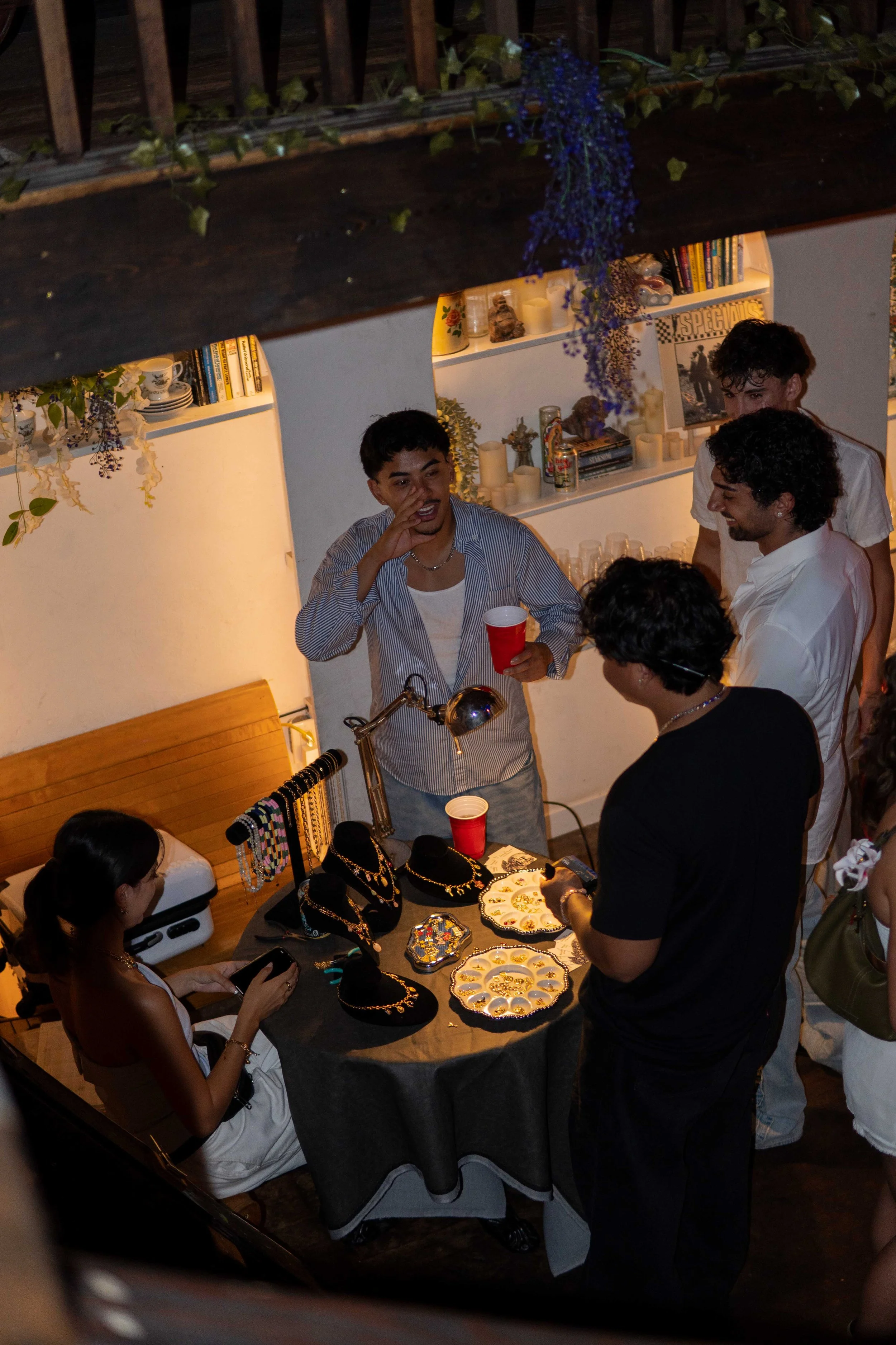 Group of people at a jewelry display table during a social gathering or party, with jewelry and decorative plates on the table, and books and candles on shelves in the background.