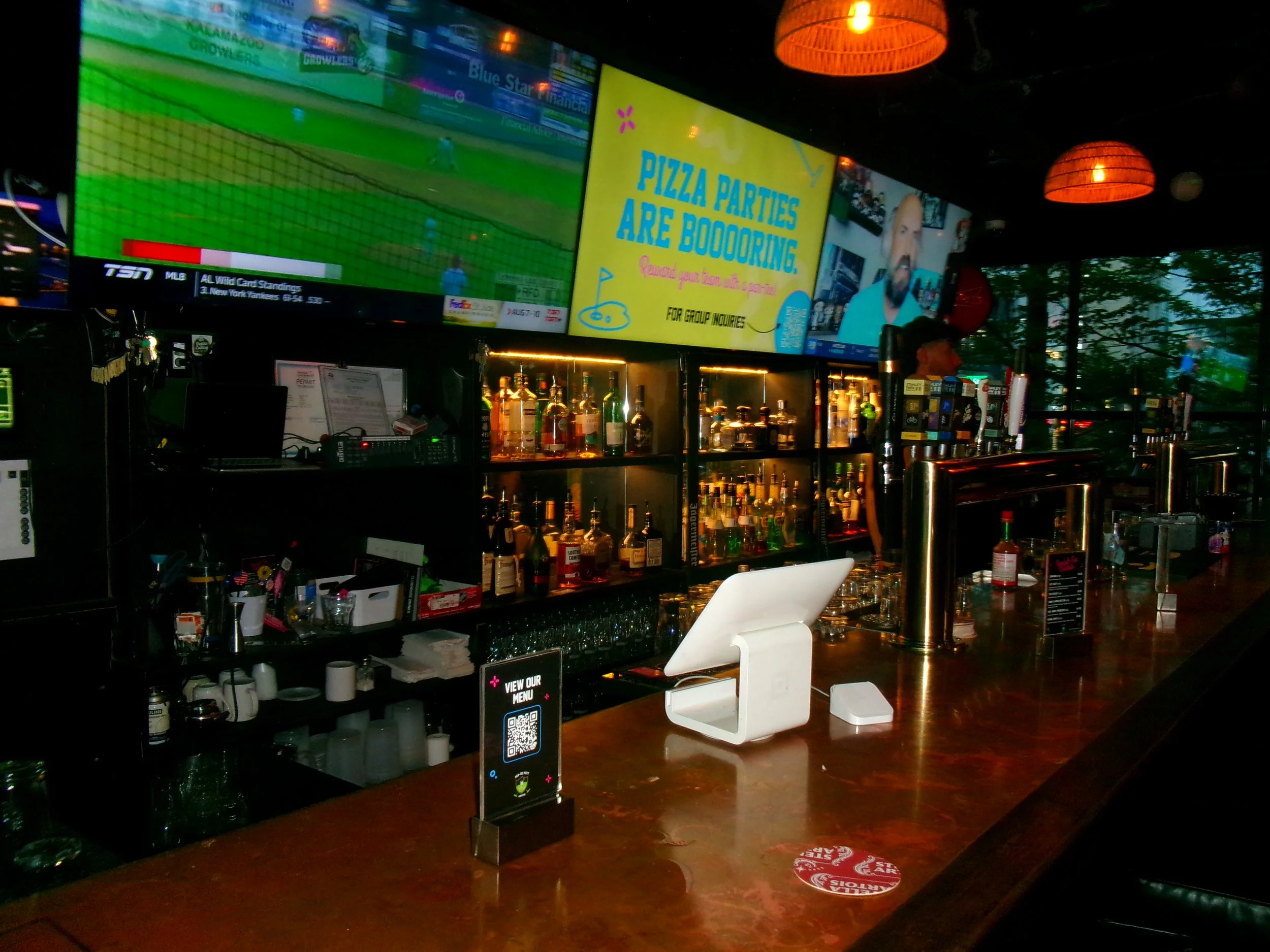 Bar with liquor bottles, TV screens showing sports, a sign advertising pizza parties, a white stand with a QR code, and a wooden counter in a dimly lit setting with orange hanging lights.