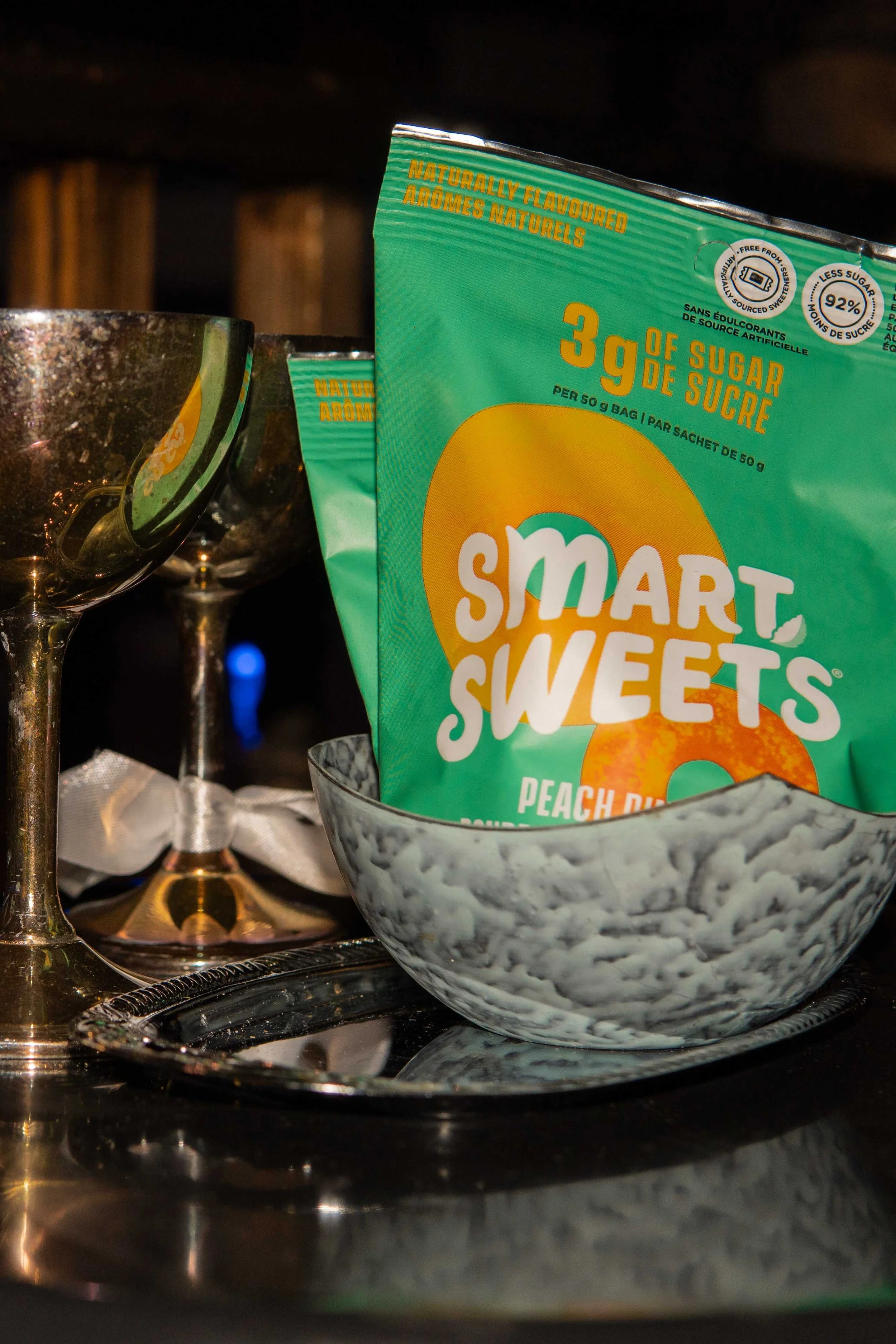 A package of Smart Sweets peach rings in a gray bowl, flanked by two empty metallic cocktail glasses, on a reflective tray with a napkin and dark background.