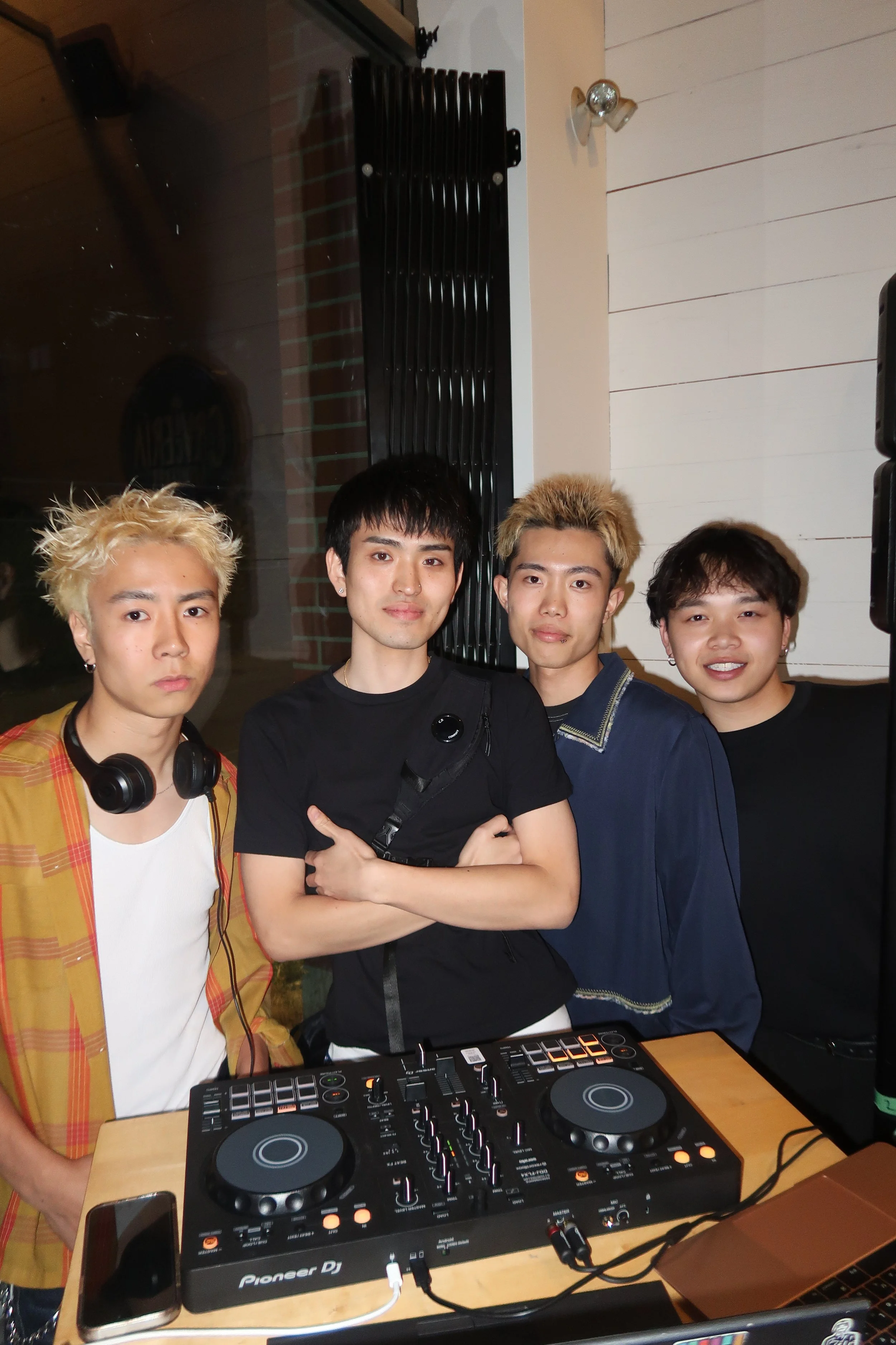 Four young men posing behind DJ equipment. One wears headphones around his neck, and others stand close, smiling at the camera, in a room with a microphone and some sound equipment.
