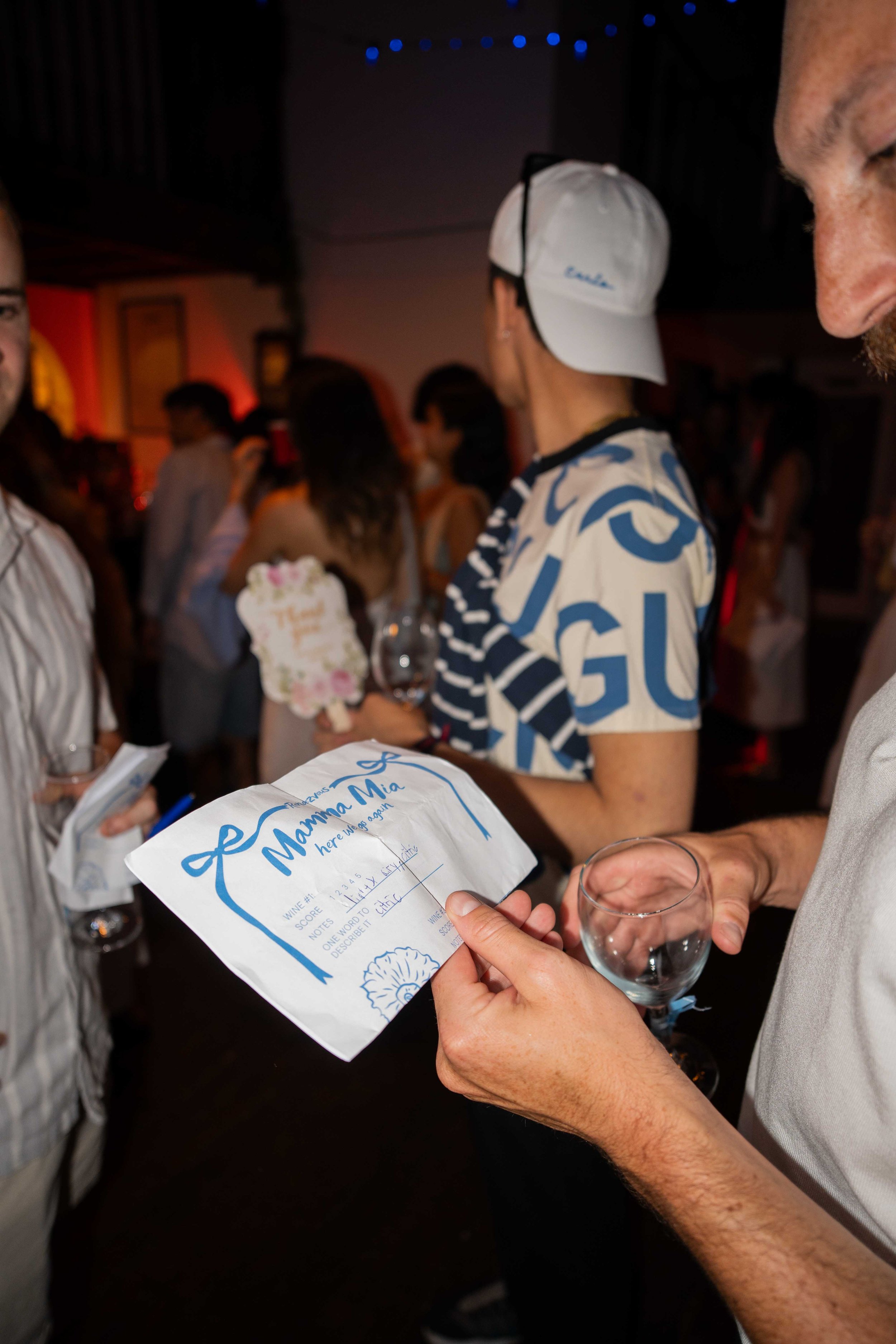 Person holding a certificate for a wine tasting event, with a group of people in the background at a social gathering or party.