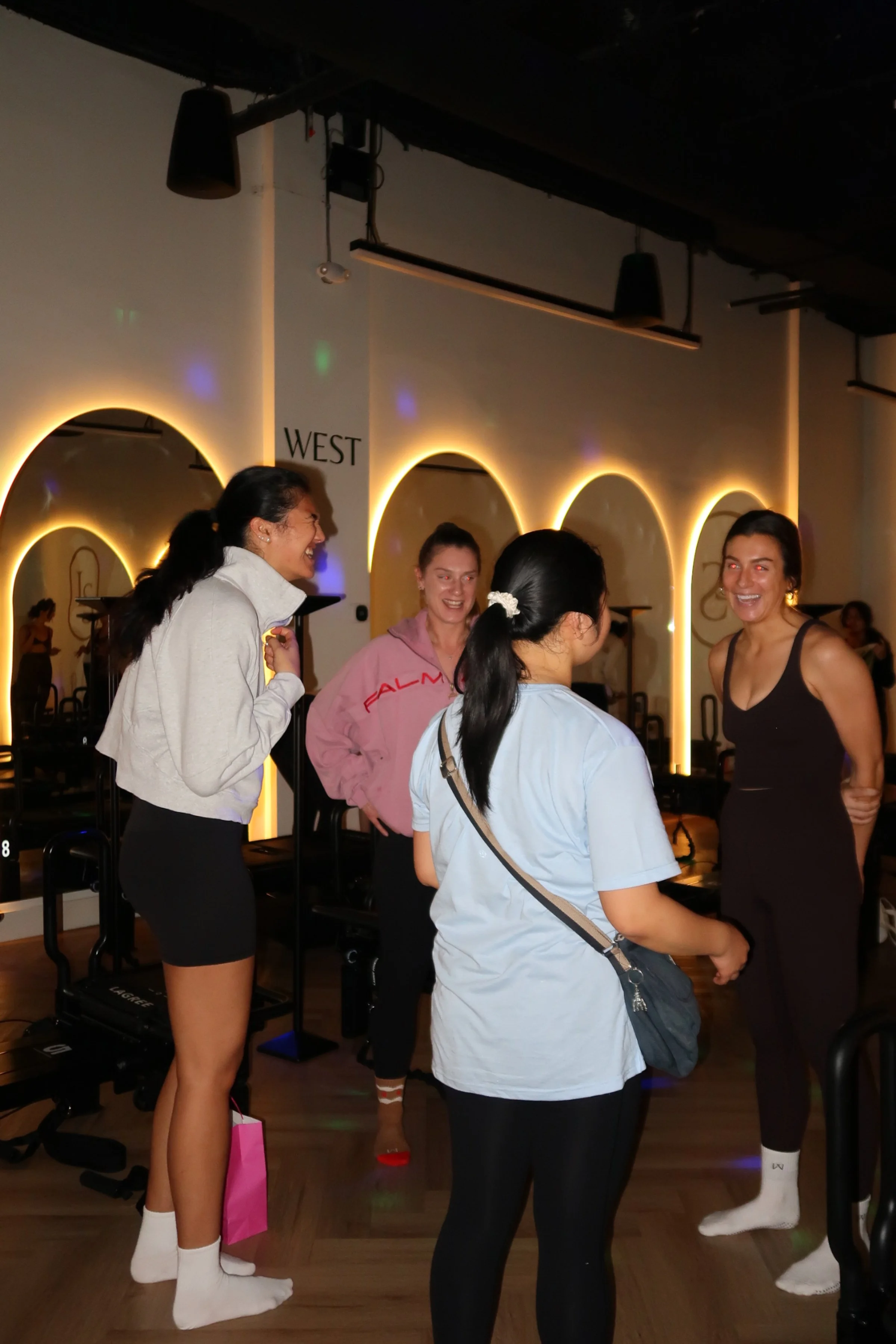 Four women in a fitness studio, engaging in conversation, some smiling, with a mirrored wall and glowing neon arches in the background.