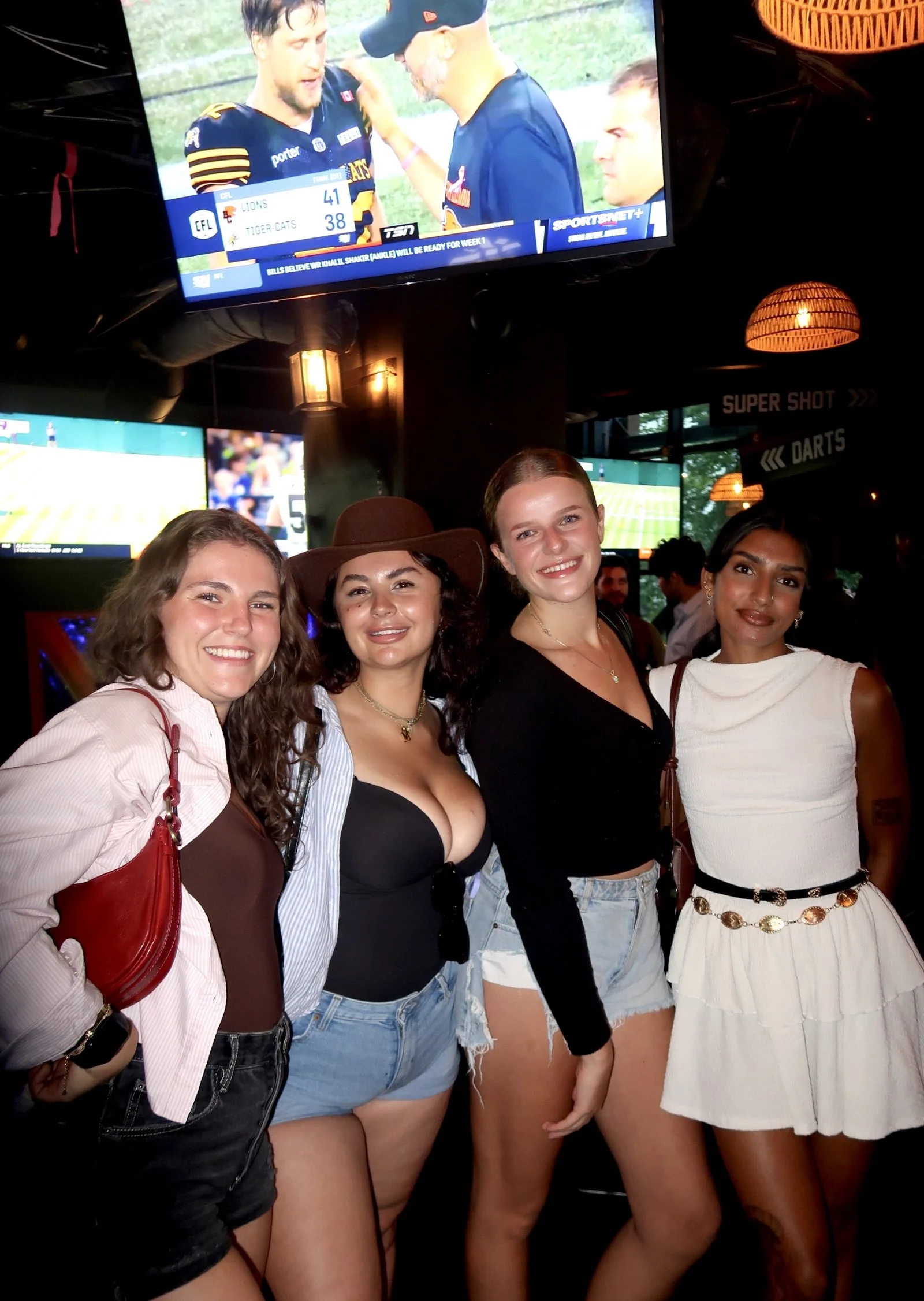 Four women posing together inside a sports bar or restaurant, with multiple TV screens displaying sports games and scores in the background.