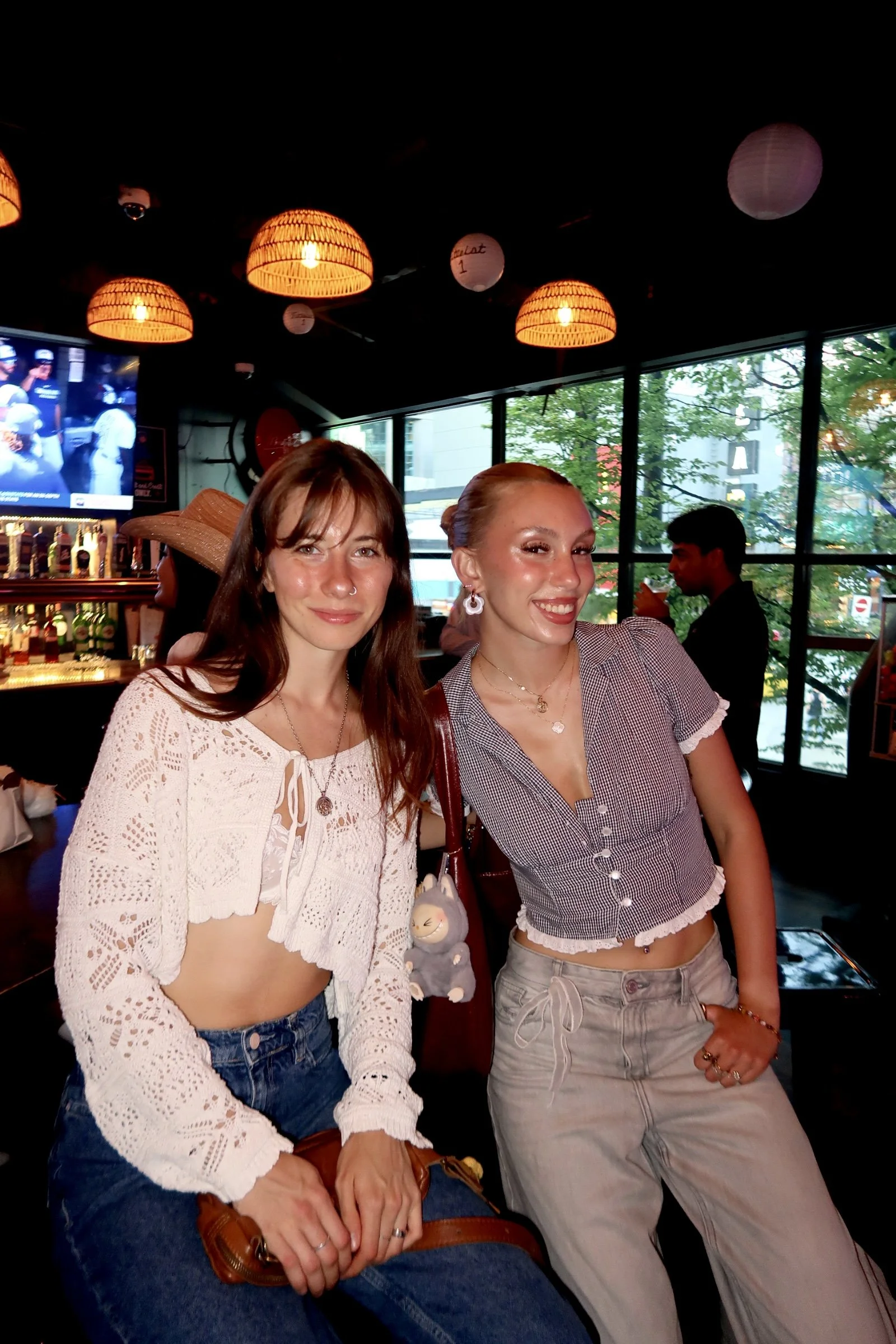 Two young women posing and smiling inside a bar or restaurant with hanging lights, large windows showing trees outside, and a TV screen in the background.