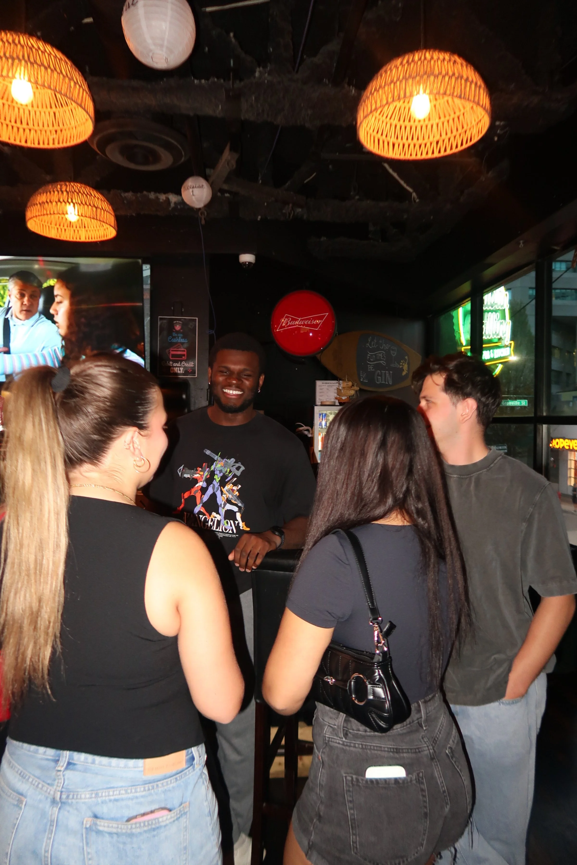 Group of people talking and laughing inside a bar or pub with orange hanging lights and neon signs on the wall
