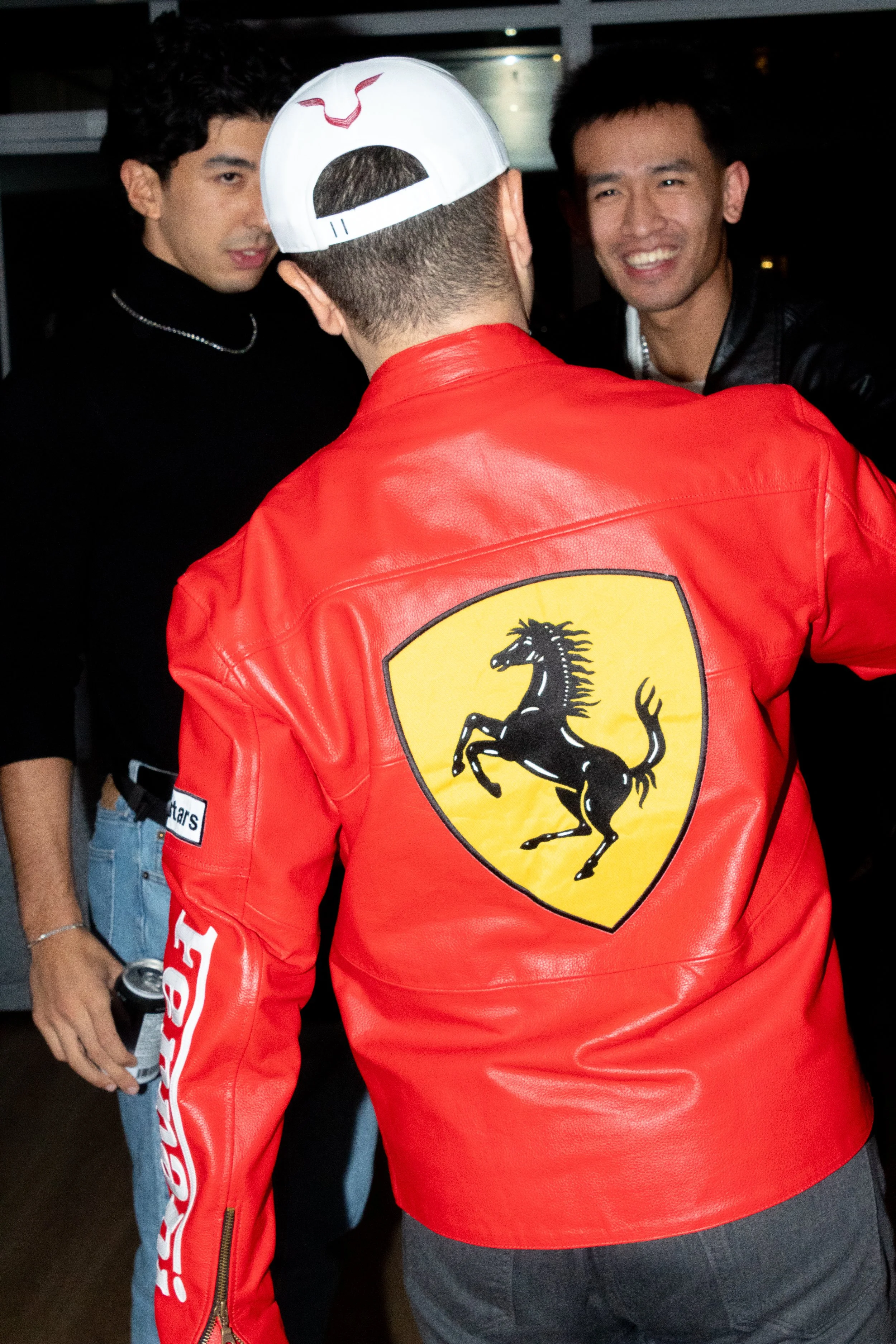 Three young men socializing, one wearing a red Ferrari leather jacket with a black prancing horse logo on the back and a white cap, others in dark clothing.