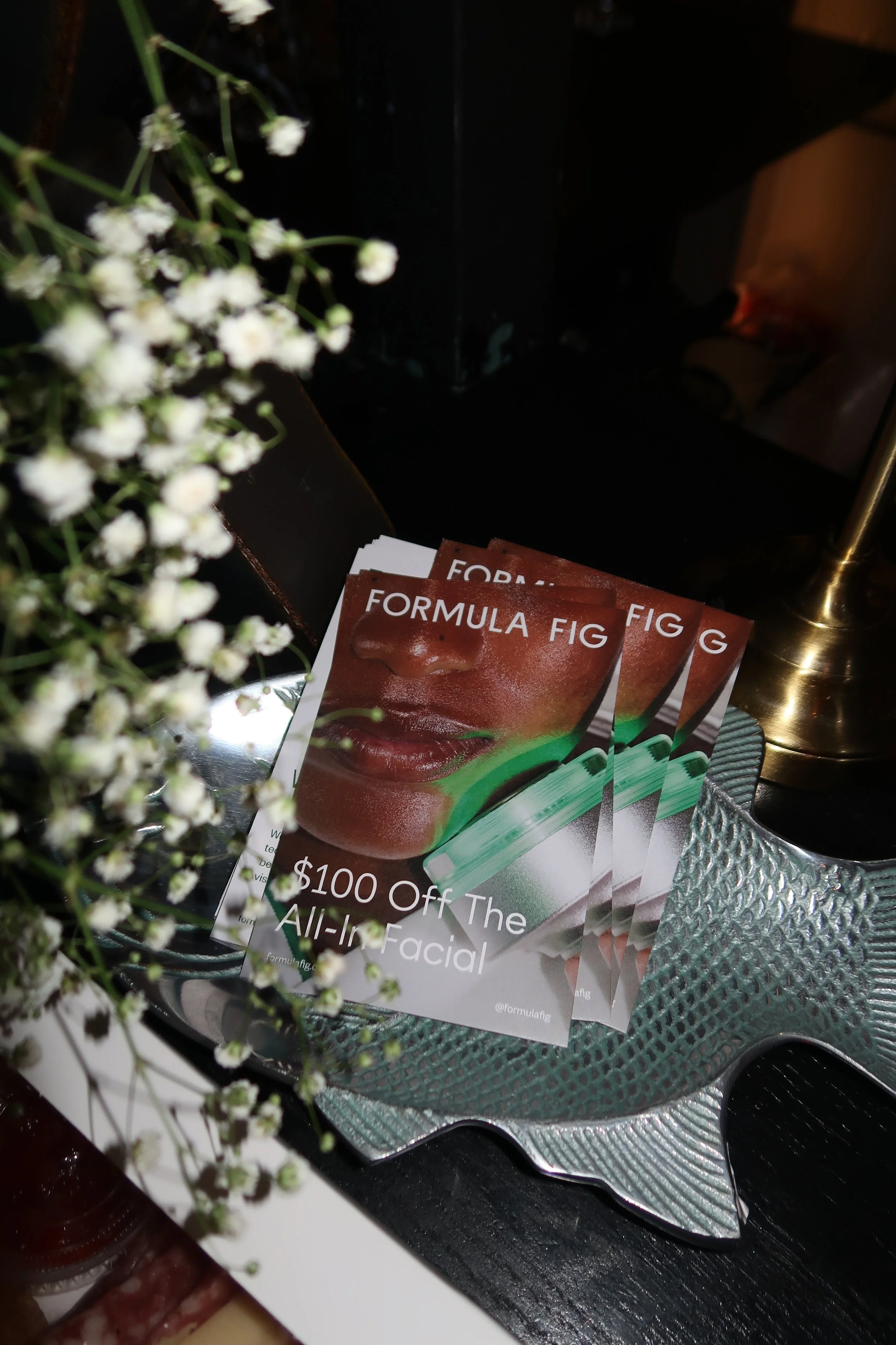 Promotional cards for Formula Fig, offering $100 off the all-in facial, with a close-up of a person's face and lips and some white flowers partially obscuring the view.