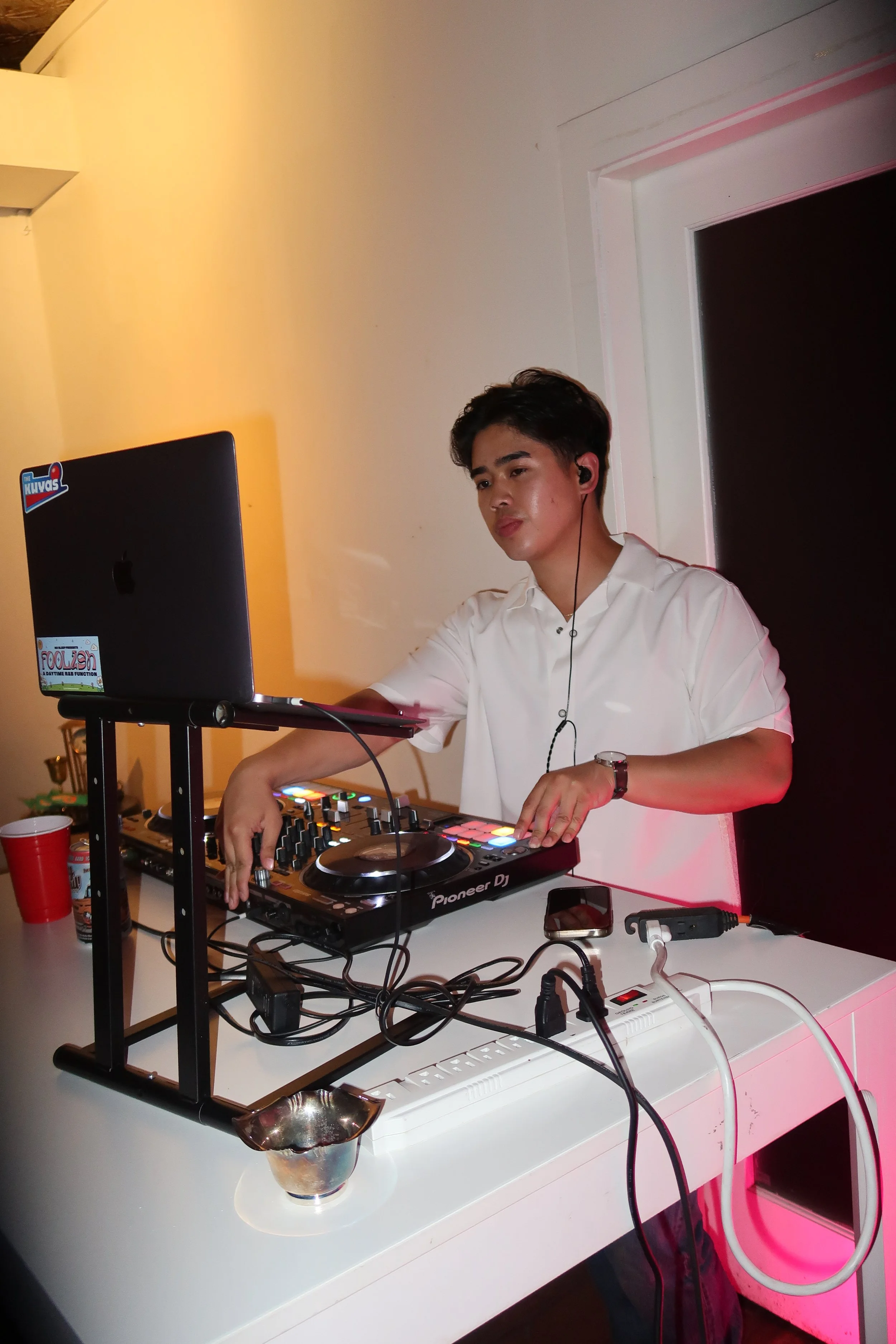 A young man in a white shirt DJing with a Pioneer DJ controller, a laptop, and a smartphone on a white table, with various cables and a paper cup nearby.