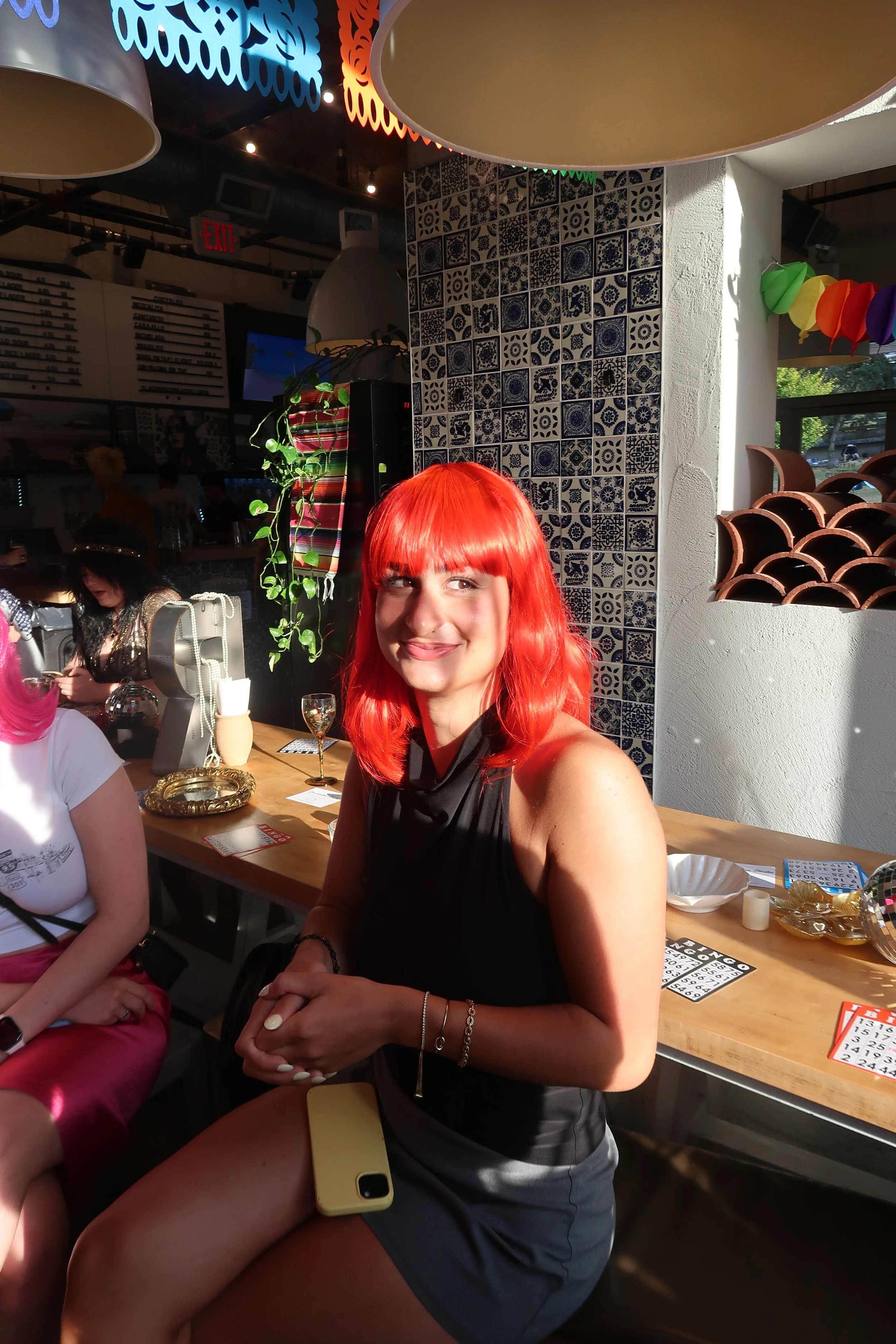 A woman with bright red hair and a black sleeveless top sitting at a bar or restaurant counter, smiling, with sunlight illuminating her face. There are bingo cards and drinks on the counter and other people in the background.