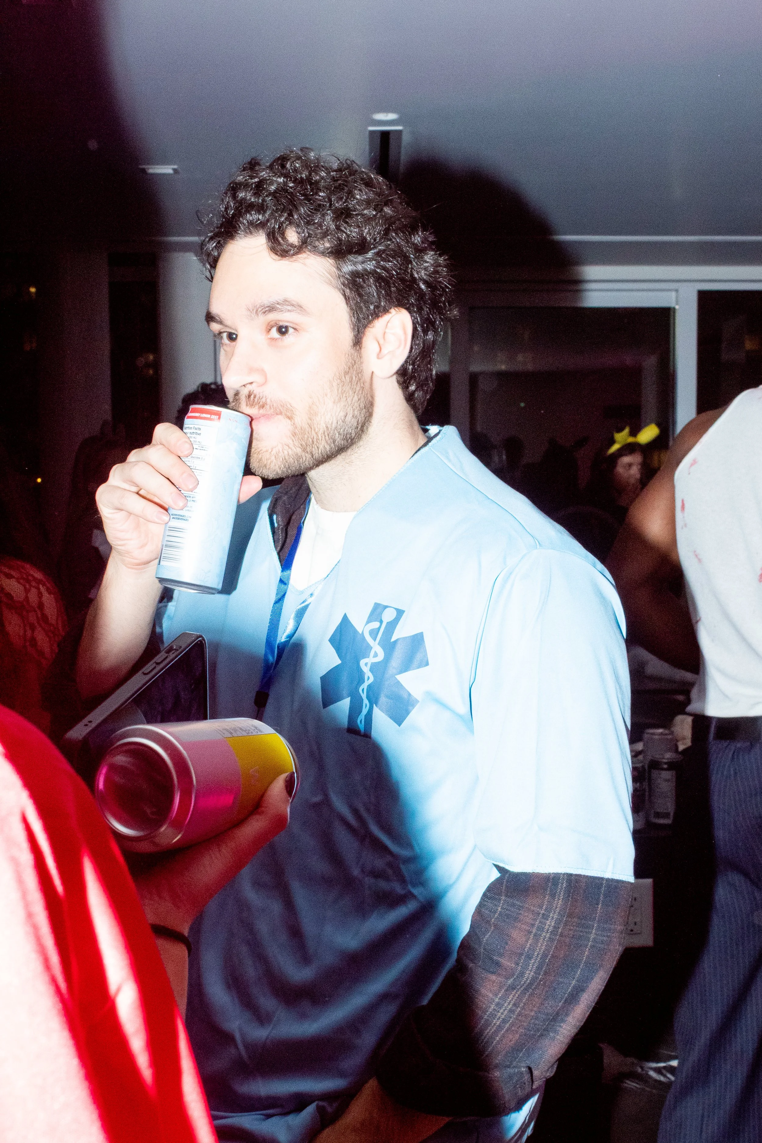 A young man with curly dark hair and a beard wearing a light blue medical shirt, holding a soda can and standing at a social gathering in an indoor setting.