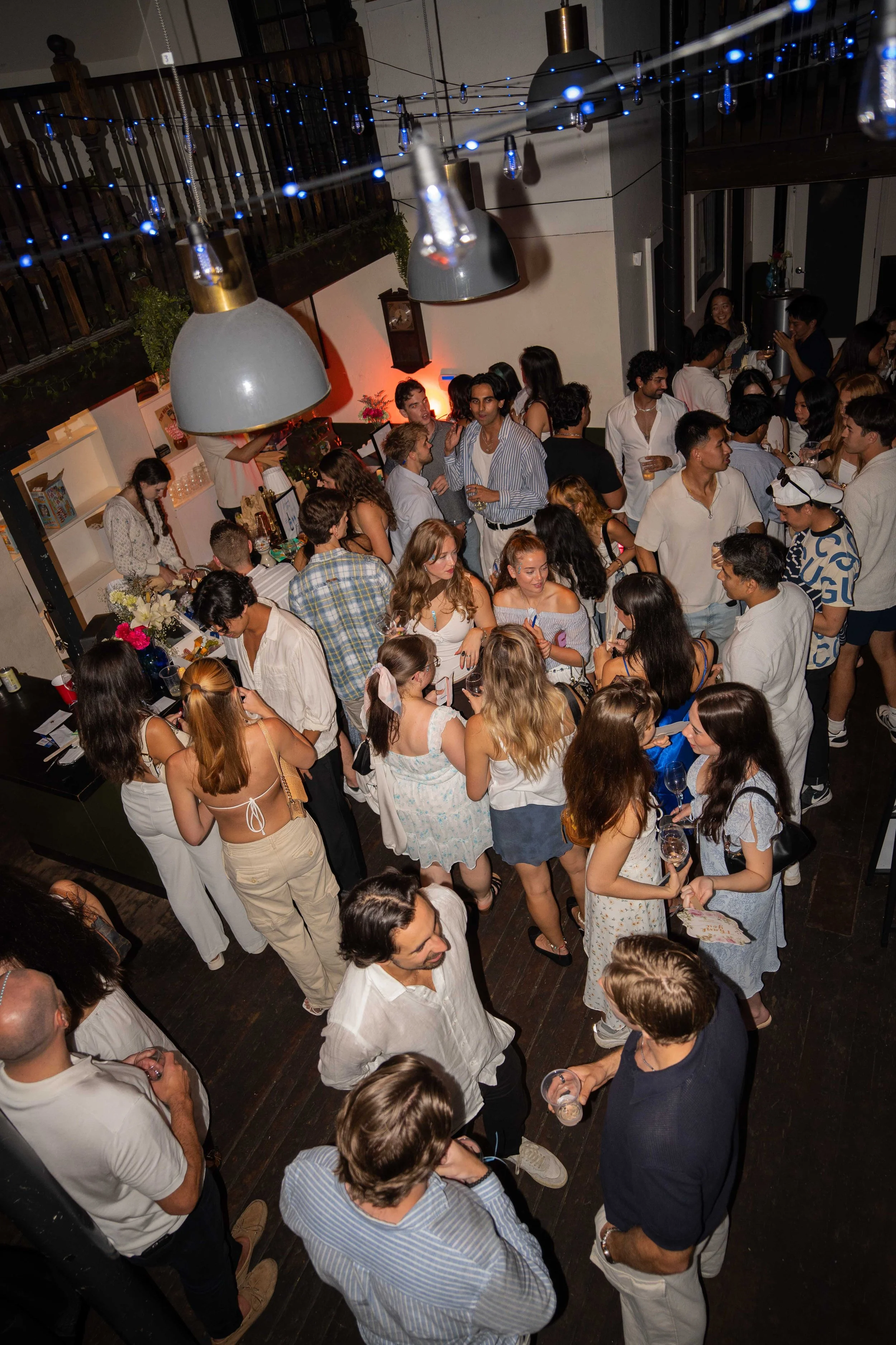 People gathered at a party inside a dimly lit venue, with some holding drinks and engaging in conversations, illuminated by overhead pendant lights, string lights, and a warm glow from the back wall.