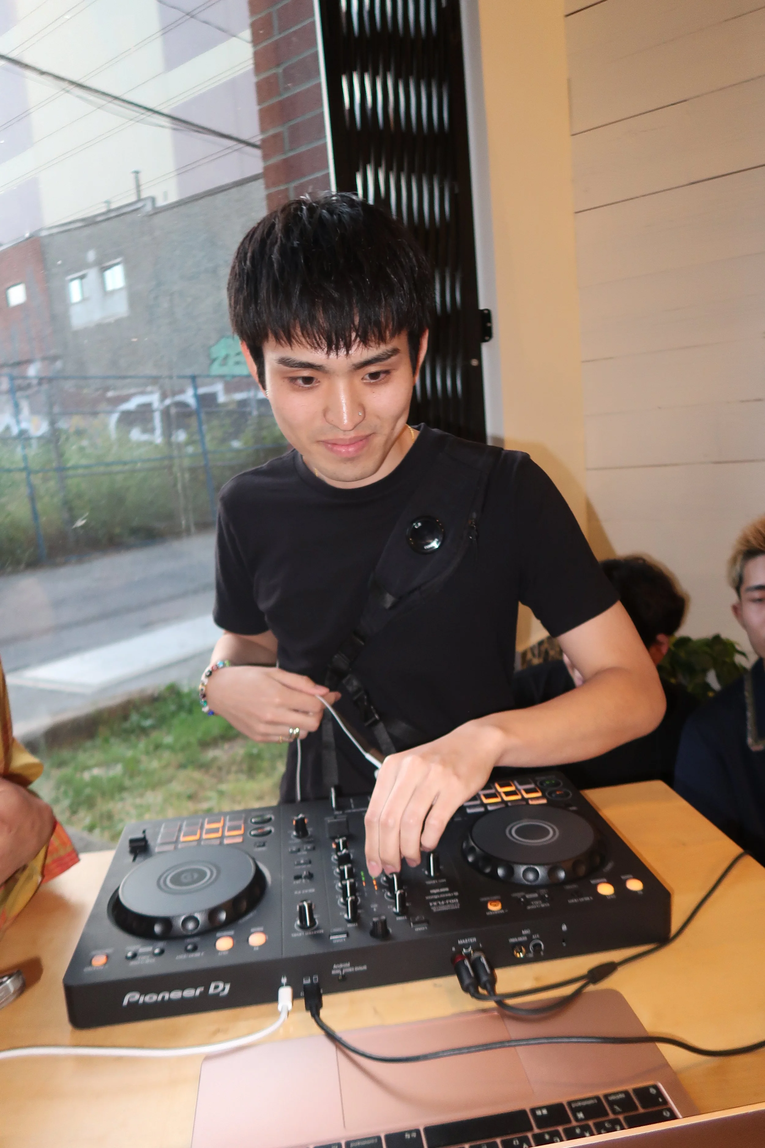 A young man with black hair and wearing a black t-shirt is operating a DJ controller at a wooden table, with a laptop nearby, indoor setting. Outside through the window, there are buildings and a fence.