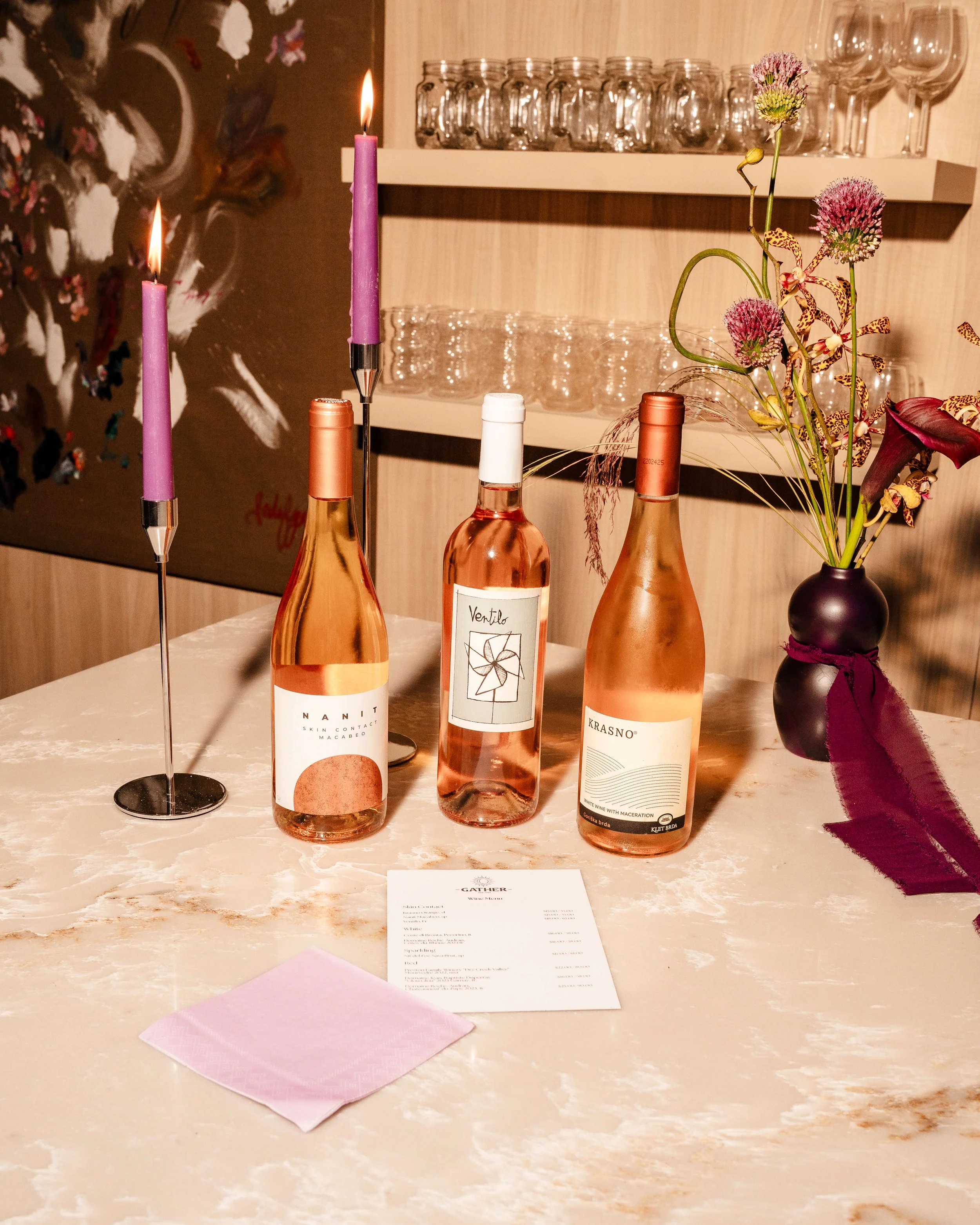 Three bottles of rosé wine, two pink candles in candle sticks, a black vase with pink and purple flowers, a pink napkin, and a printed menu on a marble table in a restaurant setting.