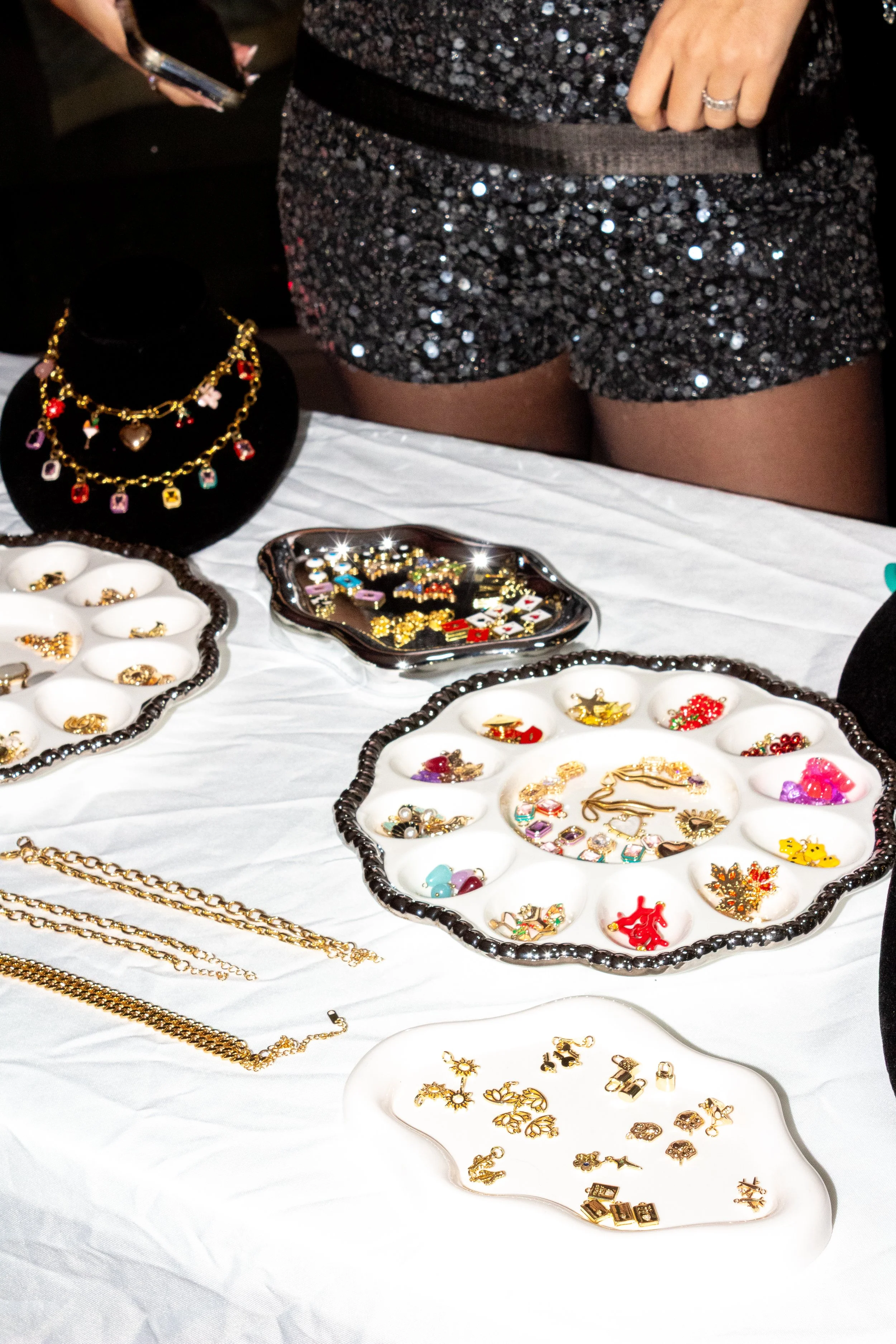 Jewelry display table with necklaces, bracelets, and earrings, including holiday-themed pins, with a person in black sparkly shorts standing nearby.