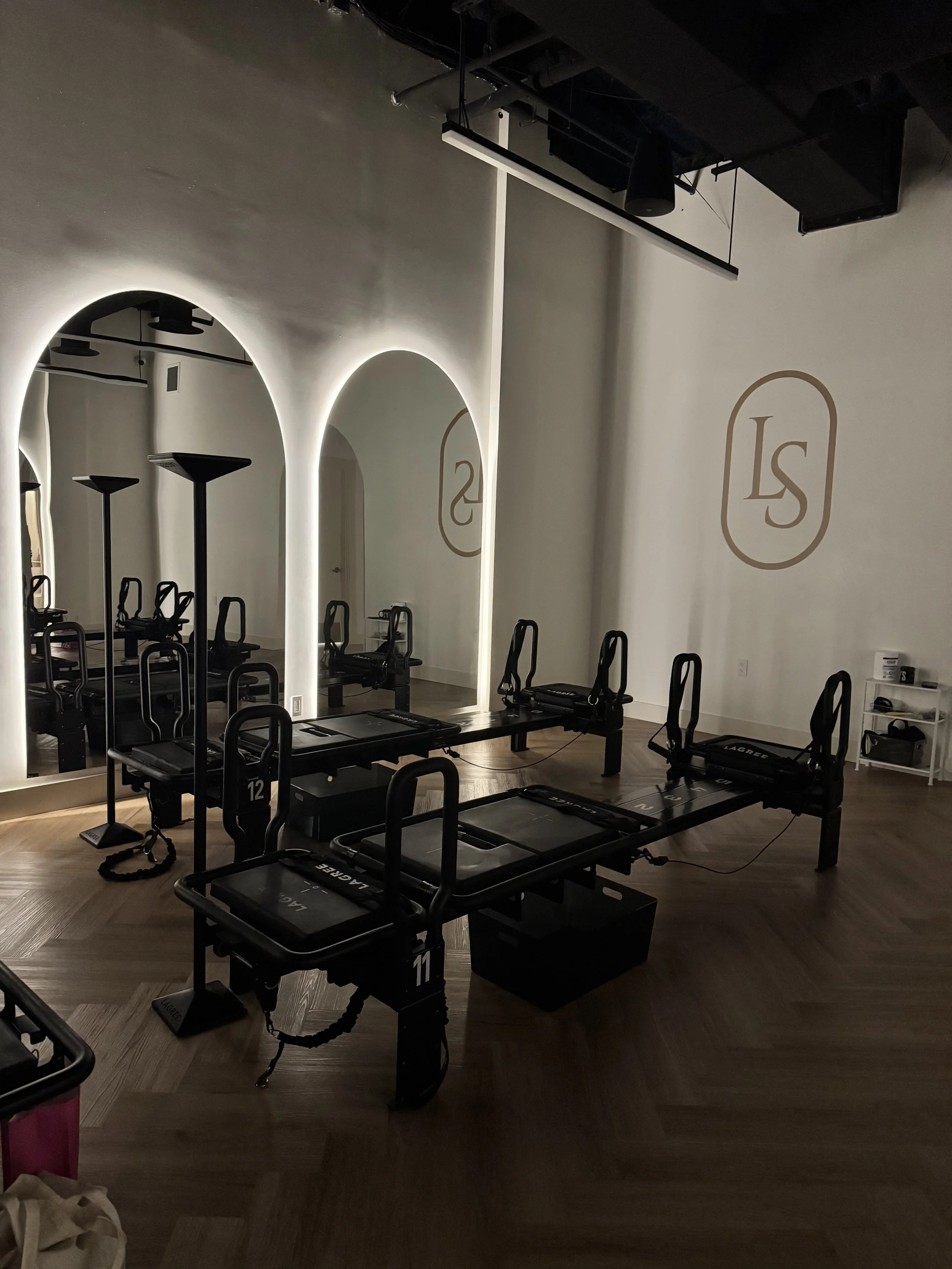 Pilates reformer machines in a dimly lit fitness studio with arched mirrors and a logo 'LS' on the wall.